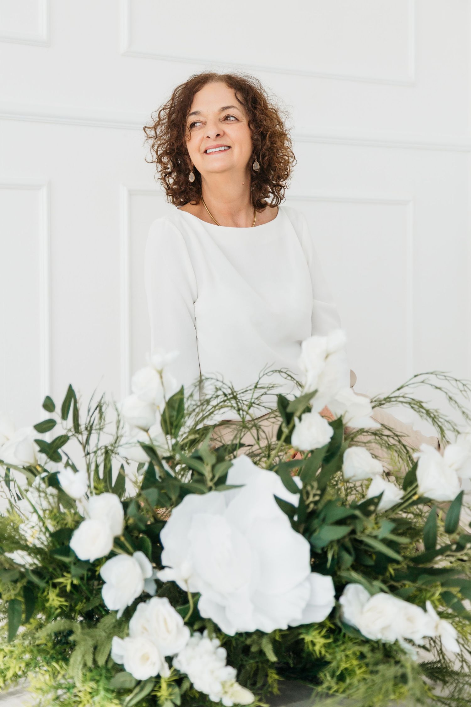 A person in a white sweater smiles while standing behind a lush arrangement of white flowers and greenery.