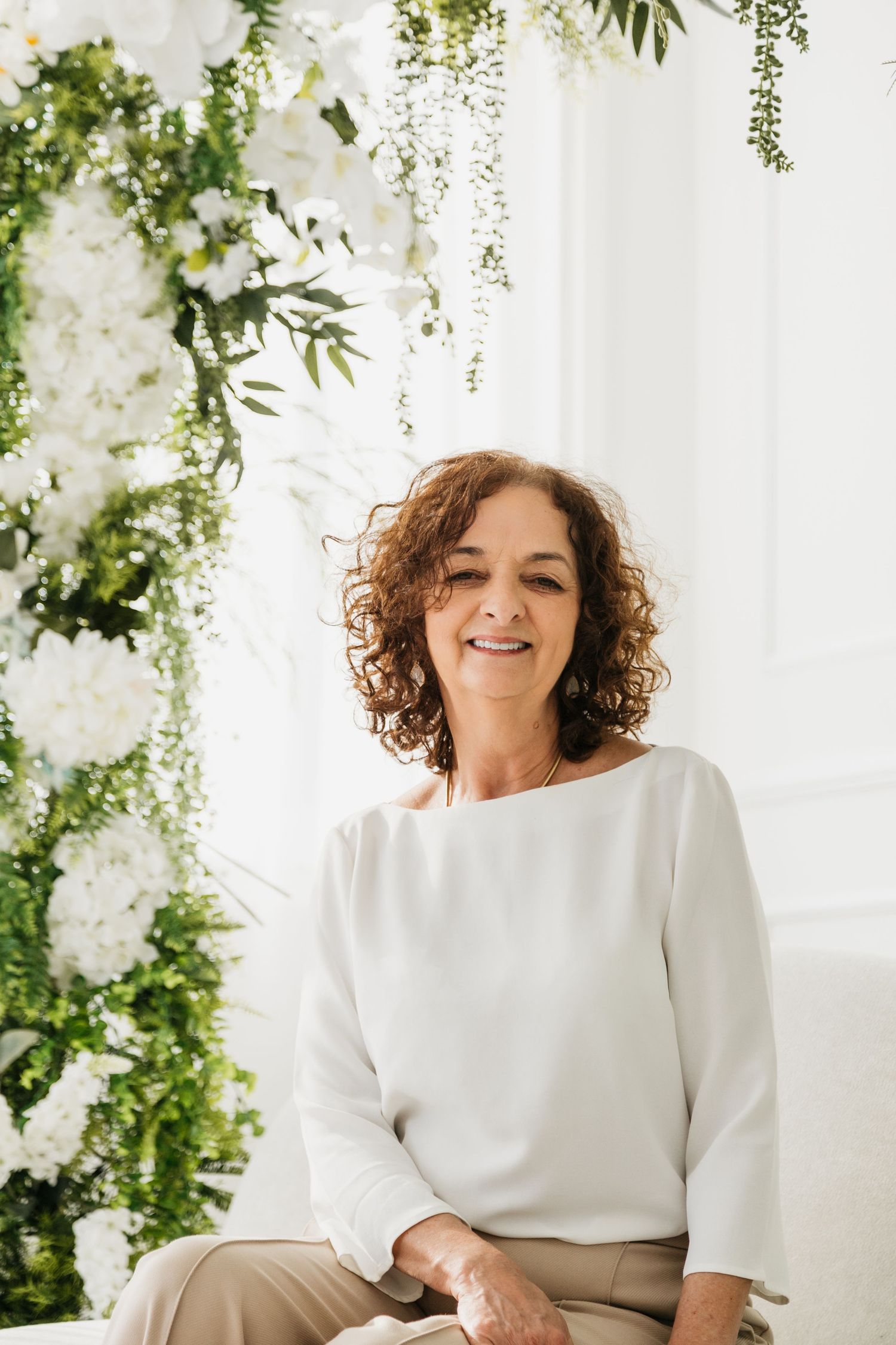 A person in a white sweater sits and smiles against a backdrop of cascading greenery and white floral decorations.