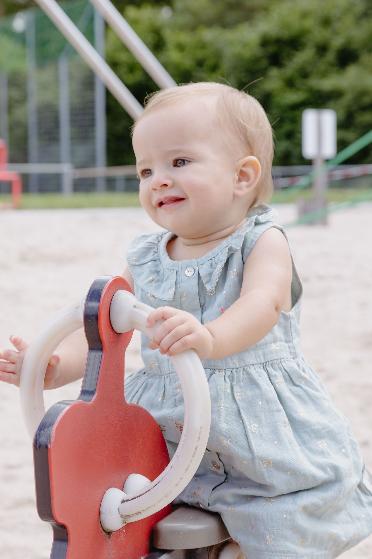 A baby in a light blue dress plays on a red spring rocker at a playground.