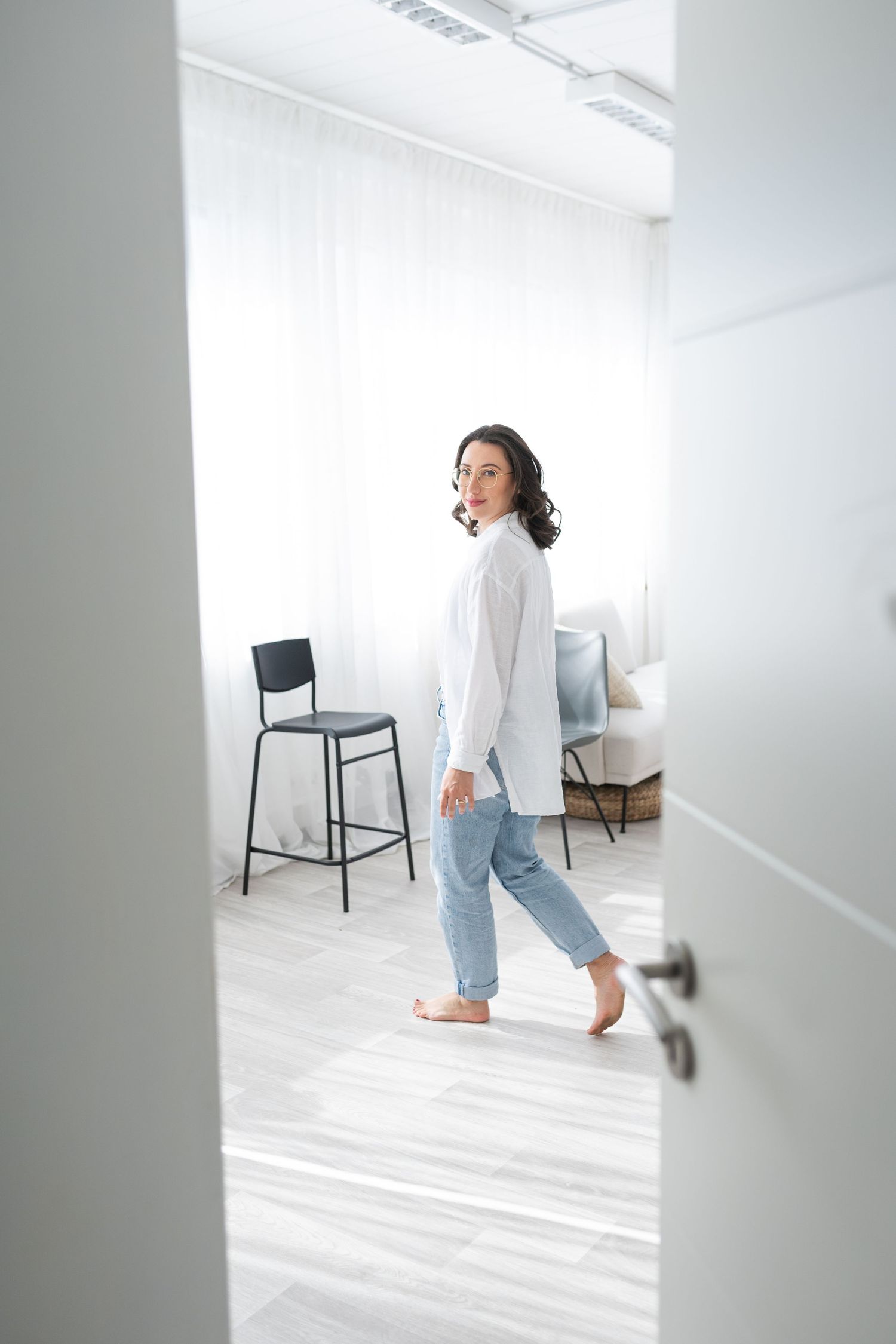 A person in a white top and light jeans walks through a minimalist white studio space with a black chair.