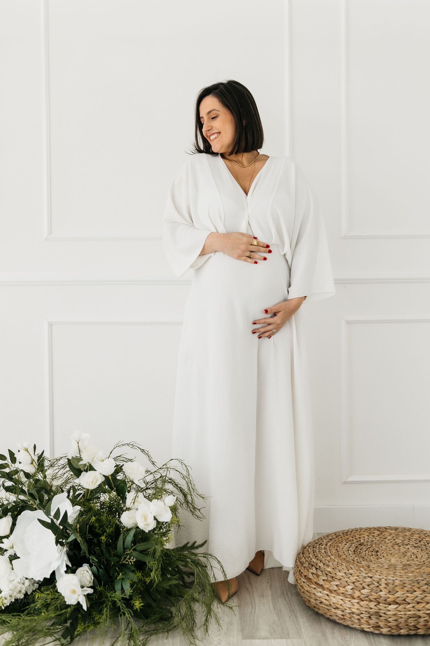 Elegant maternity photo with white flowing dress, natural decor including white florals and woven ottoman against white wall.