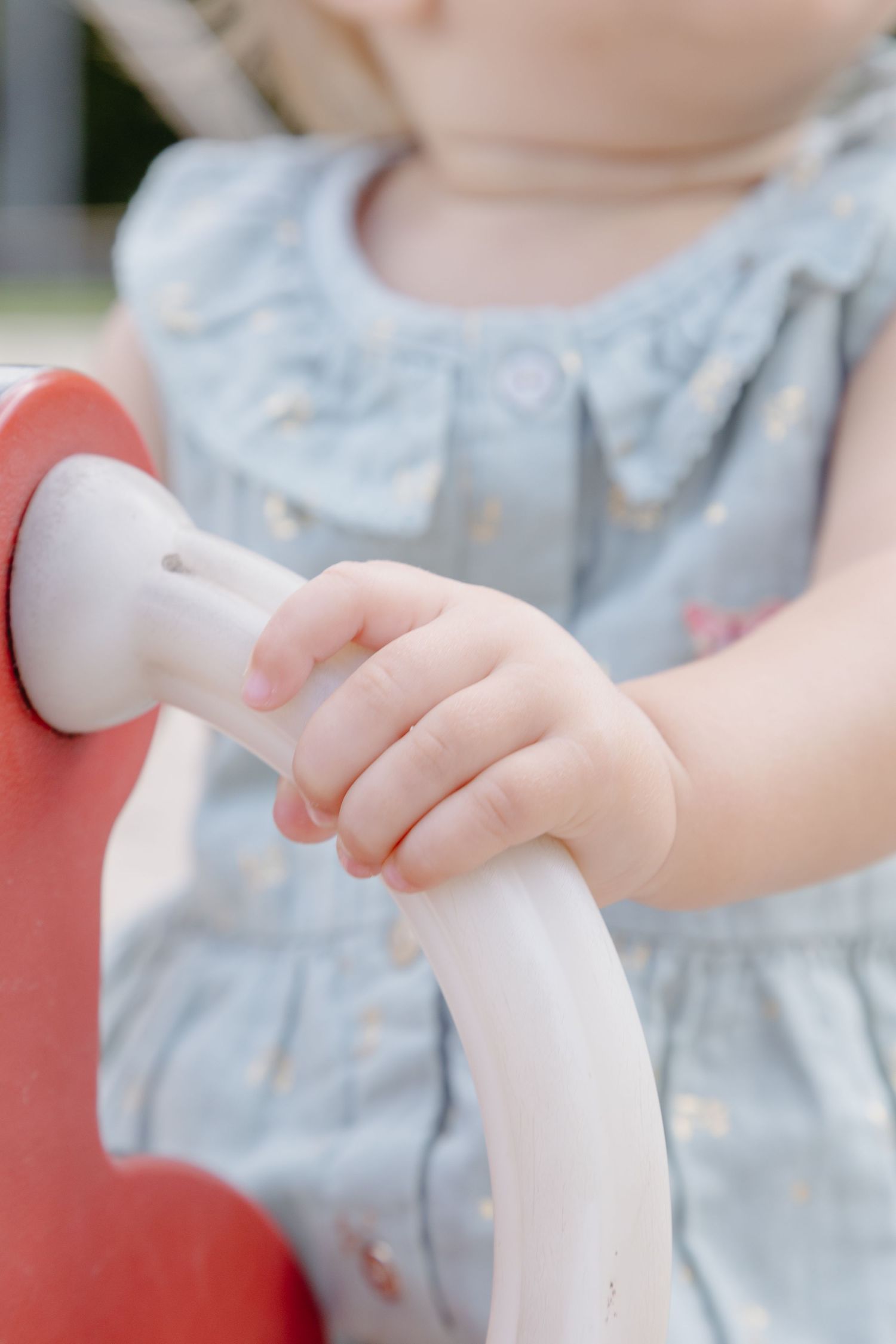 Small hands grip a white handlebar on a red playground structure while wearing a light blue dress.