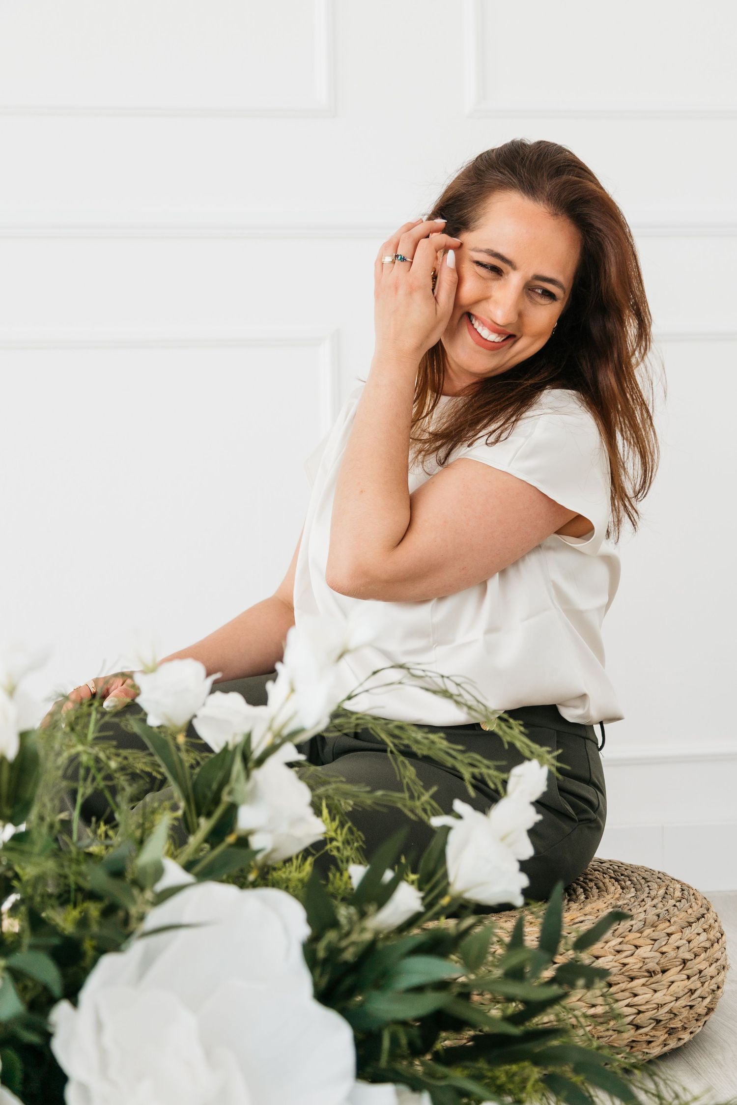 Woman in white top poses with fresh white flowers and woven straw hat against white background in lifestyle shoot.