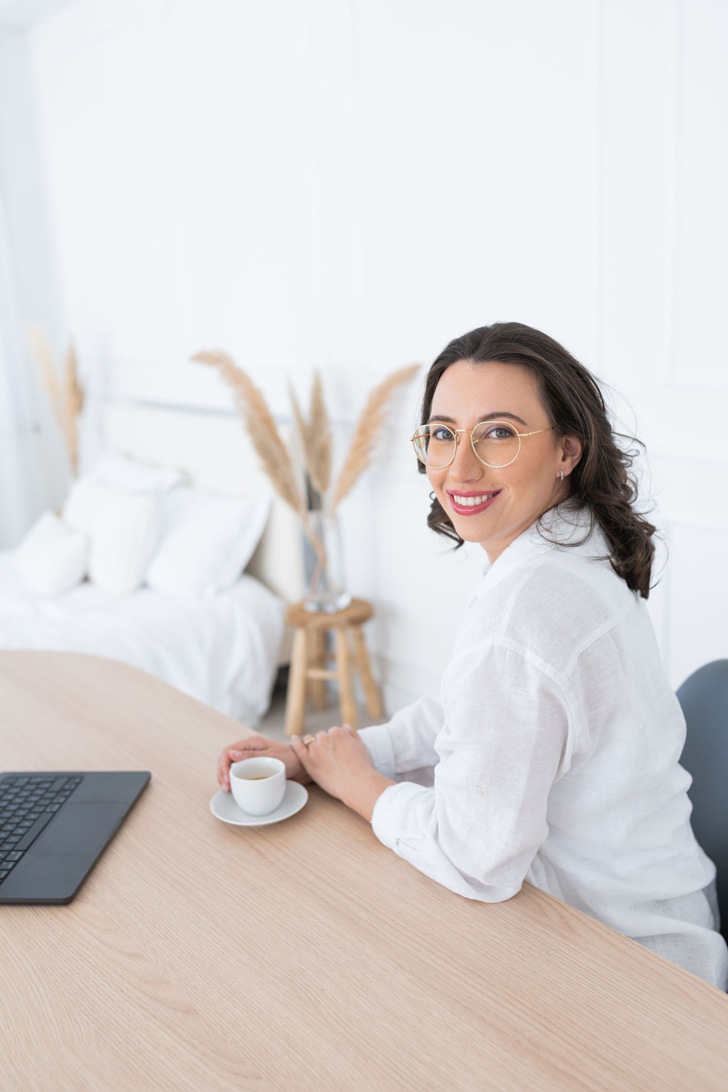 Professional wearing a white shirt and glasses sits at a wooden desk with a laptop in a bright modern workspace.