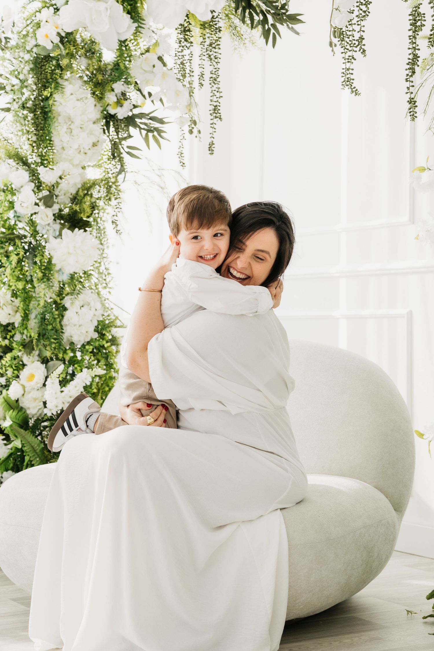 A tender moment shared between parent and child on a white couch surrounded by hanging greenery and floral decor.