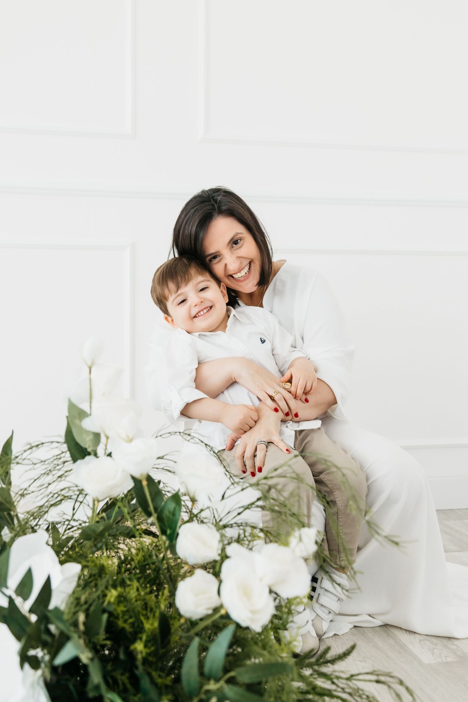 Intimate family portrait with mother and child on white backdrop surrounded by lush greenery and white floral arrangements.