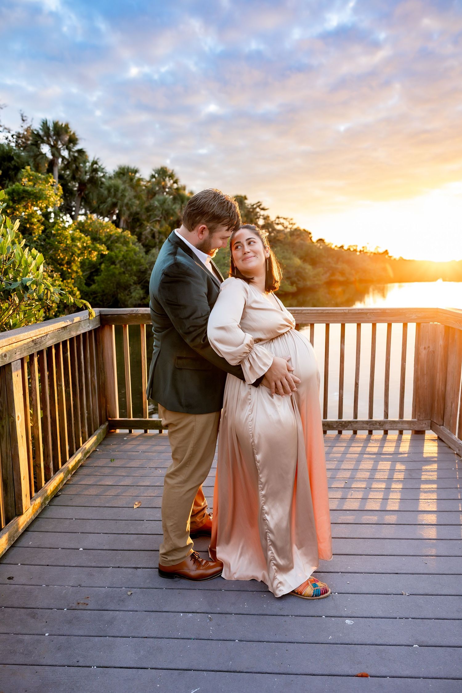 A couple shares a romantic embrace on a wooden deck overlooking a sunset river.