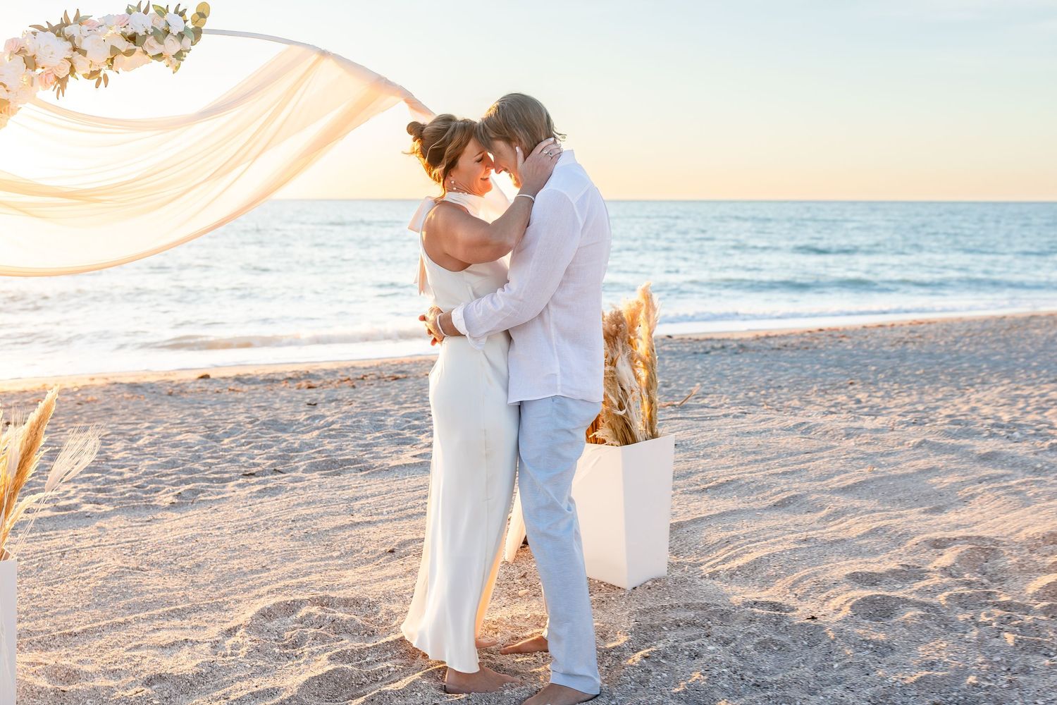 A bride and groom share their first kiss at a decorated beach wedding at sunset.