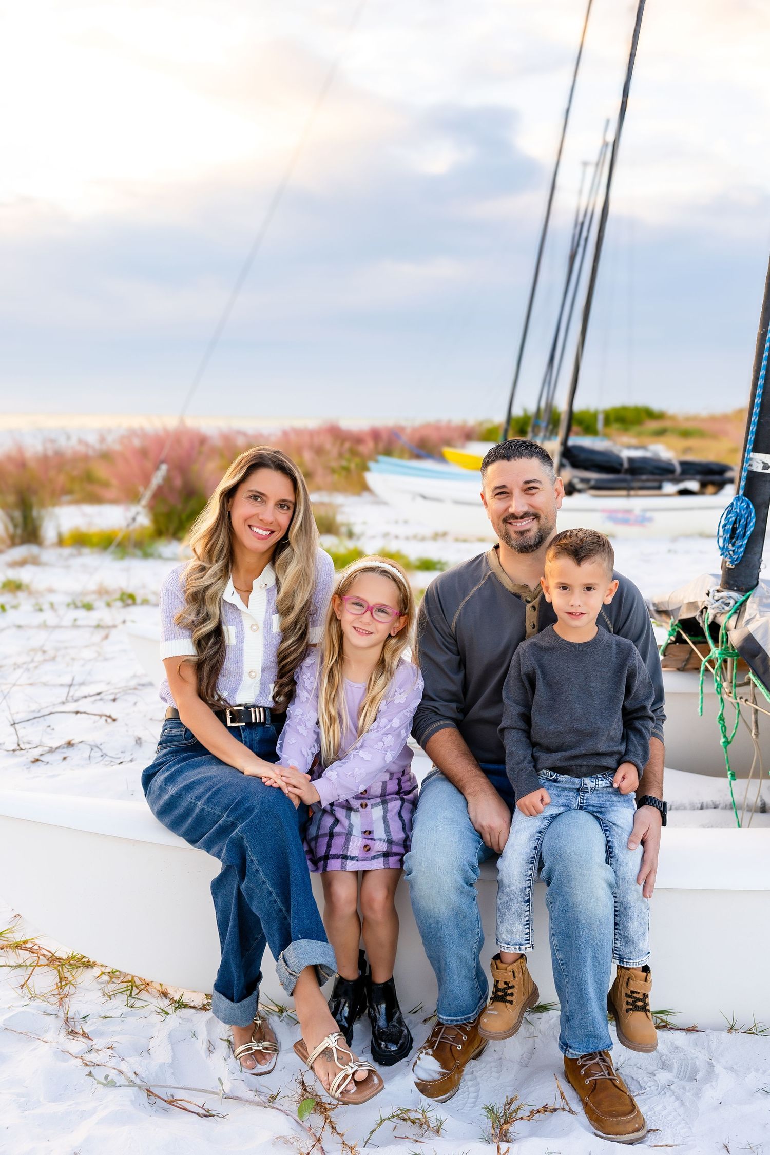 A happy family of four poses on white sand beach near a sailboat at sunset.