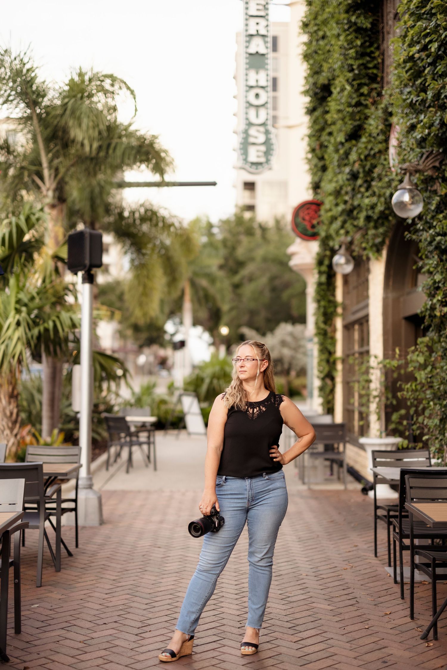 Female photographer holding camera posing on brick walkway surrounded by lush greenery and palms.
