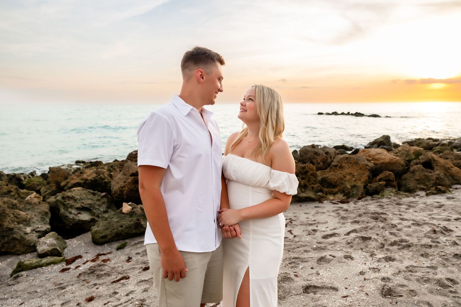 Couple embracing on a rocky beach at sunset, woman in white off-shoulder dress, man in white shirt and khakis.