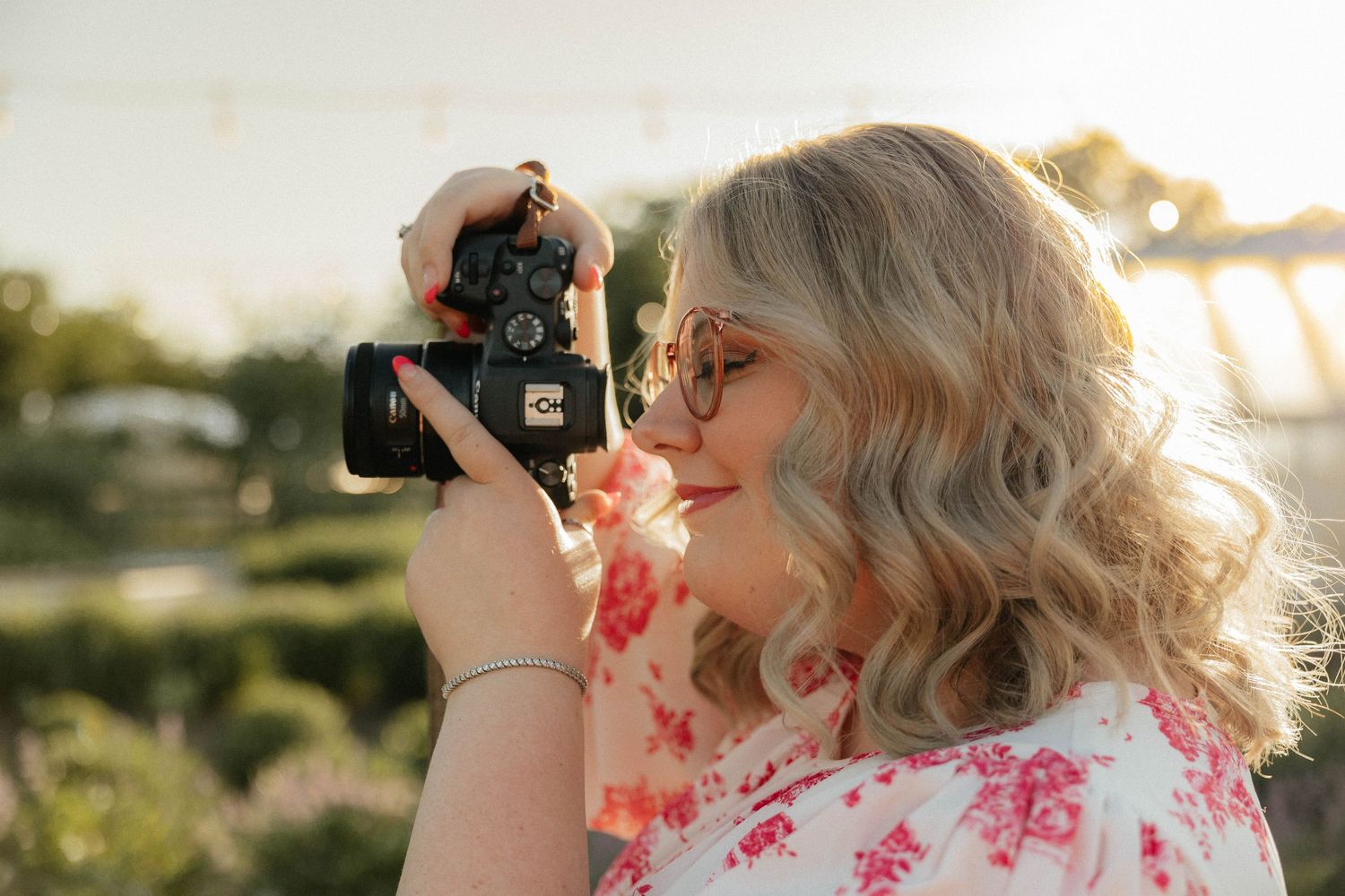 Blonde woman with glasses photographing with vintage camera in golden sunset light outdoors.