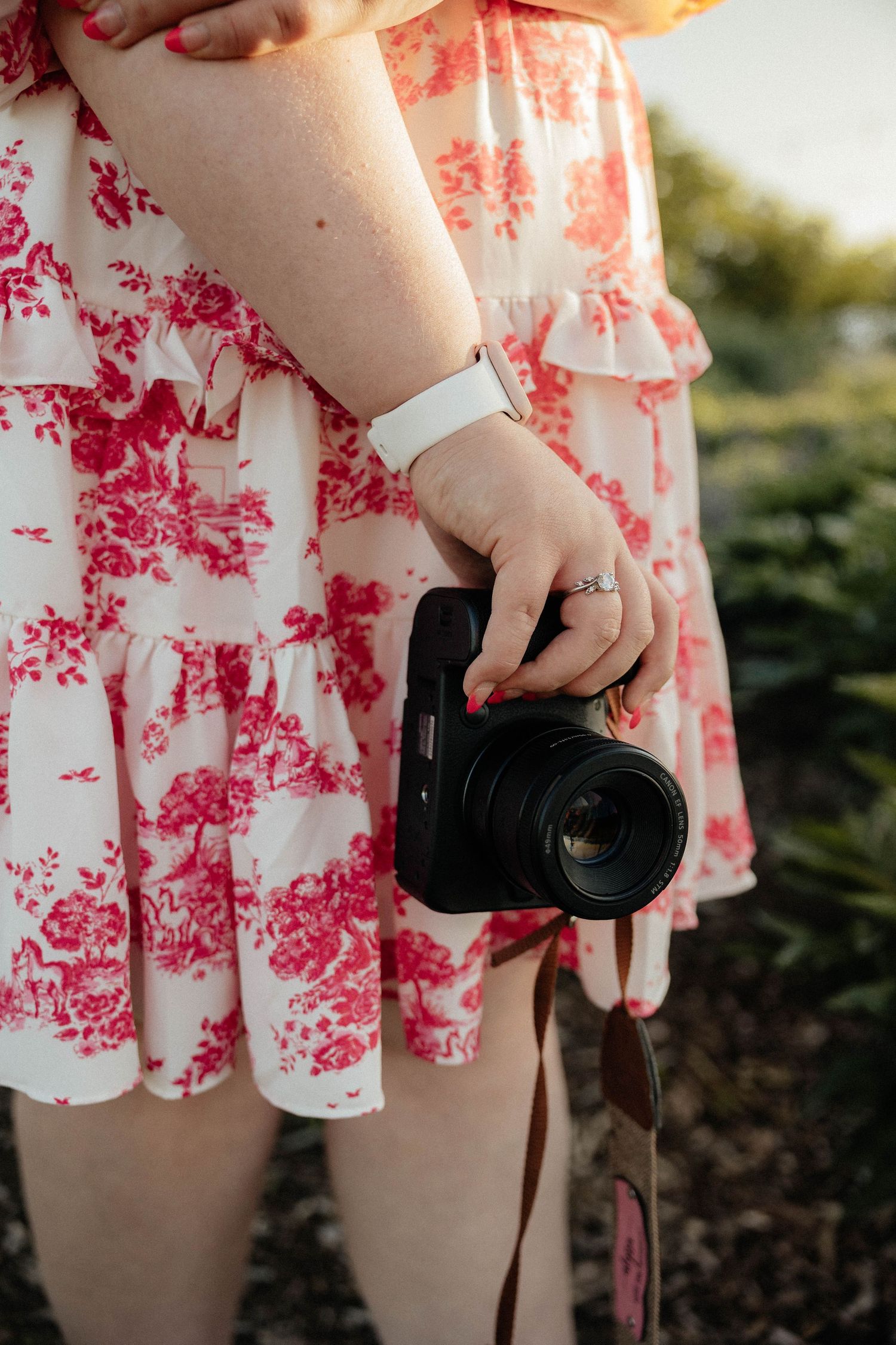 Close-up of woman in pink floral dress holding DSLR camera with ring and bracelet.
