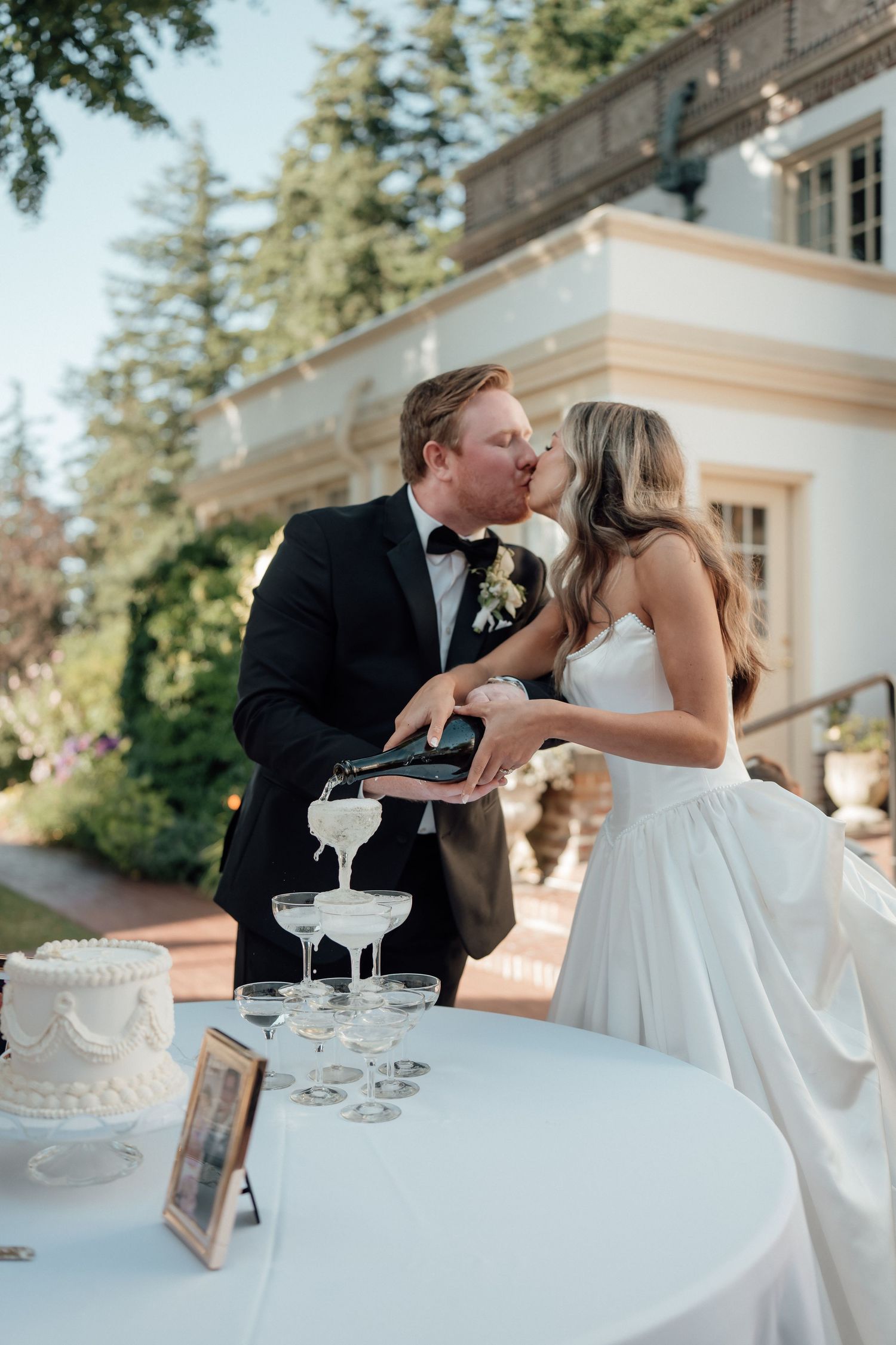 A couple pours champagne into a tower of glasses during their outdoor wedding celebration.