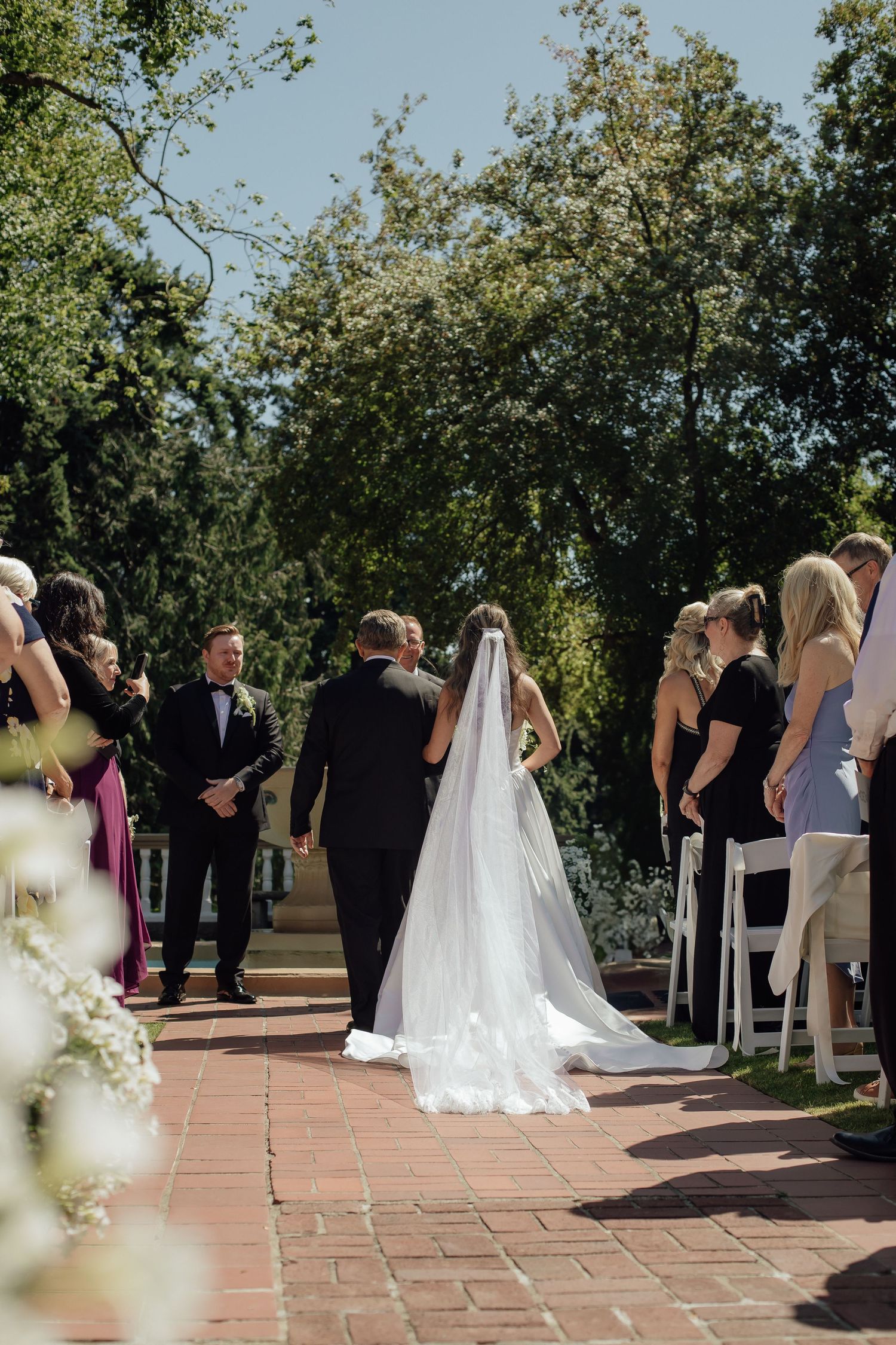 Elegant outdoor wedding ceremony with guests seated on white chairs along a brick pathway surrounded by greenery.