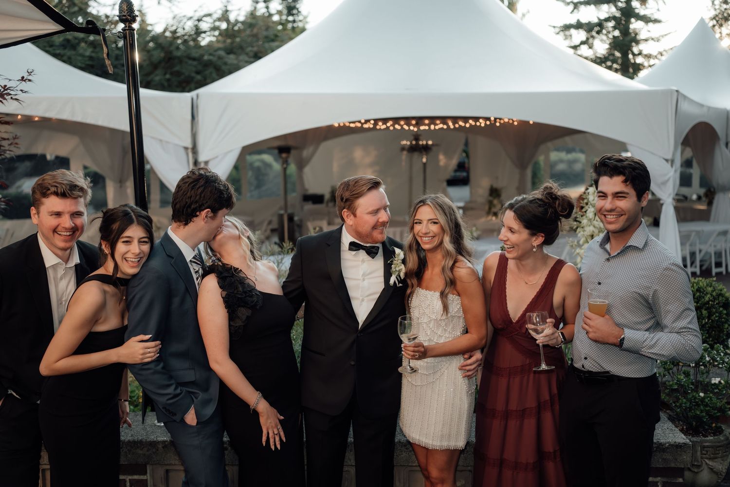 Wedding reception guests gather under white tent during sunset celebration.