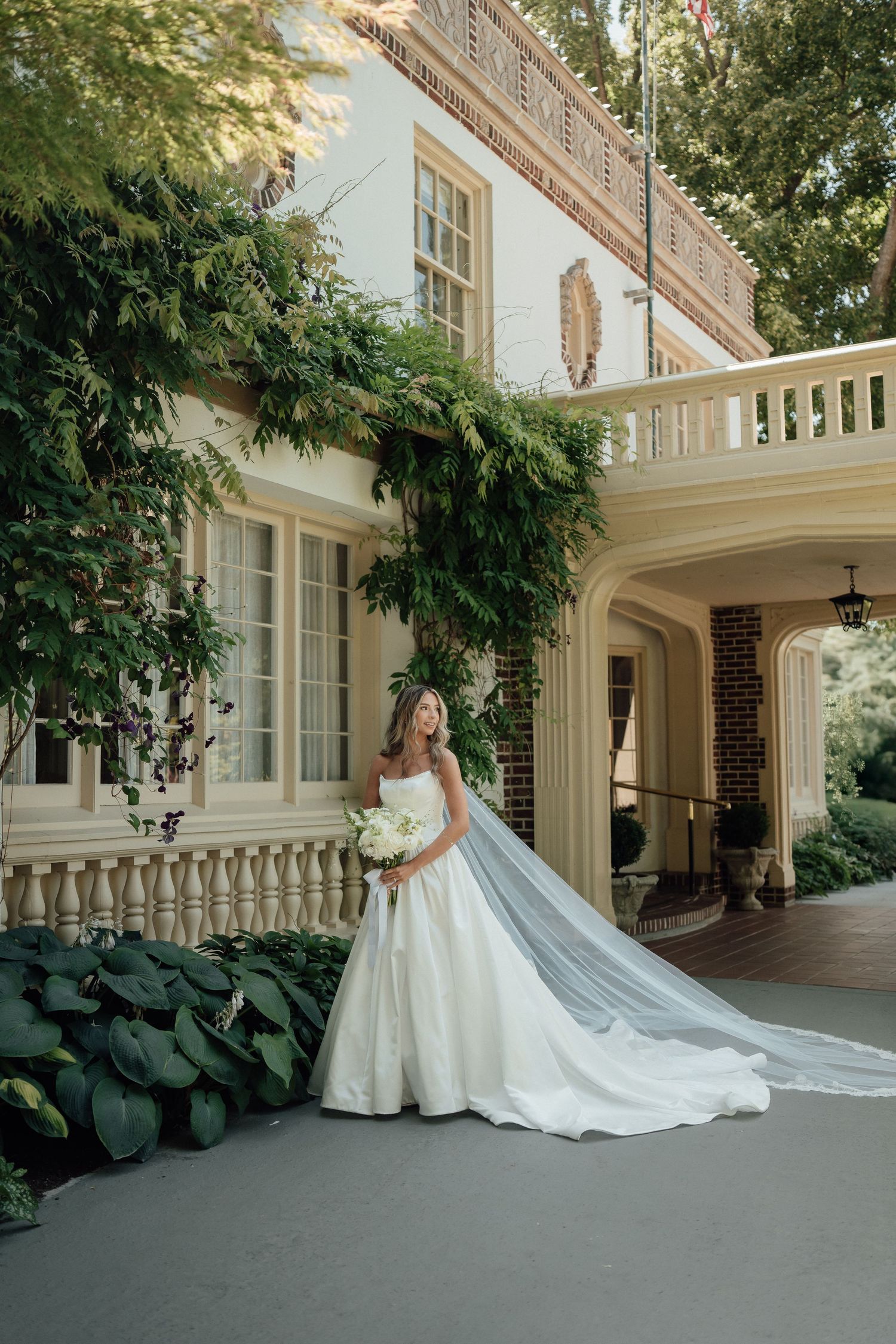 A bride in a white ballgown with cathedral veil poses in front of a grand white colonial mansion.