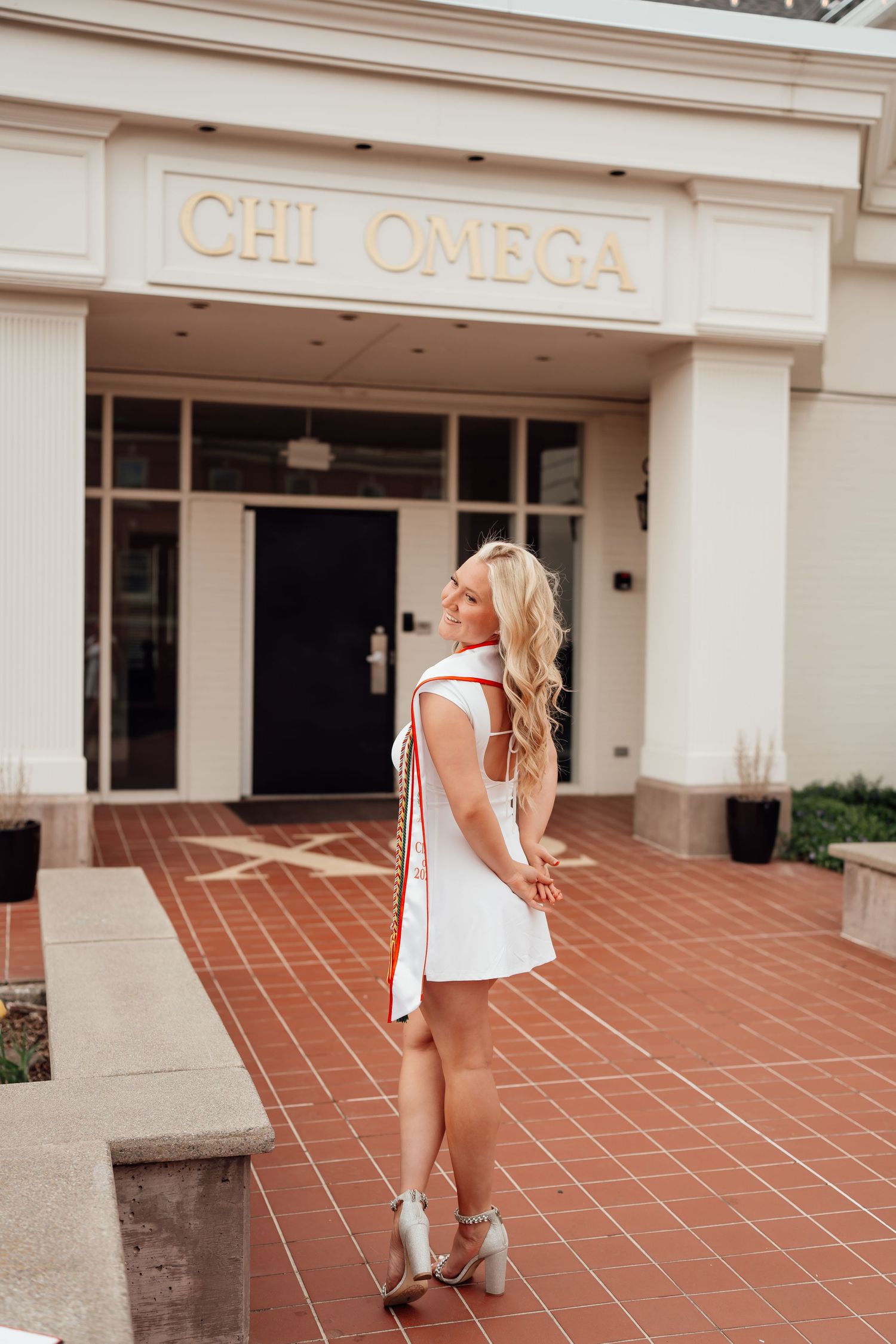Woman in white sleeveless top and shorts poses outside Chi Omega sorority house on terracotta tiled walkway.