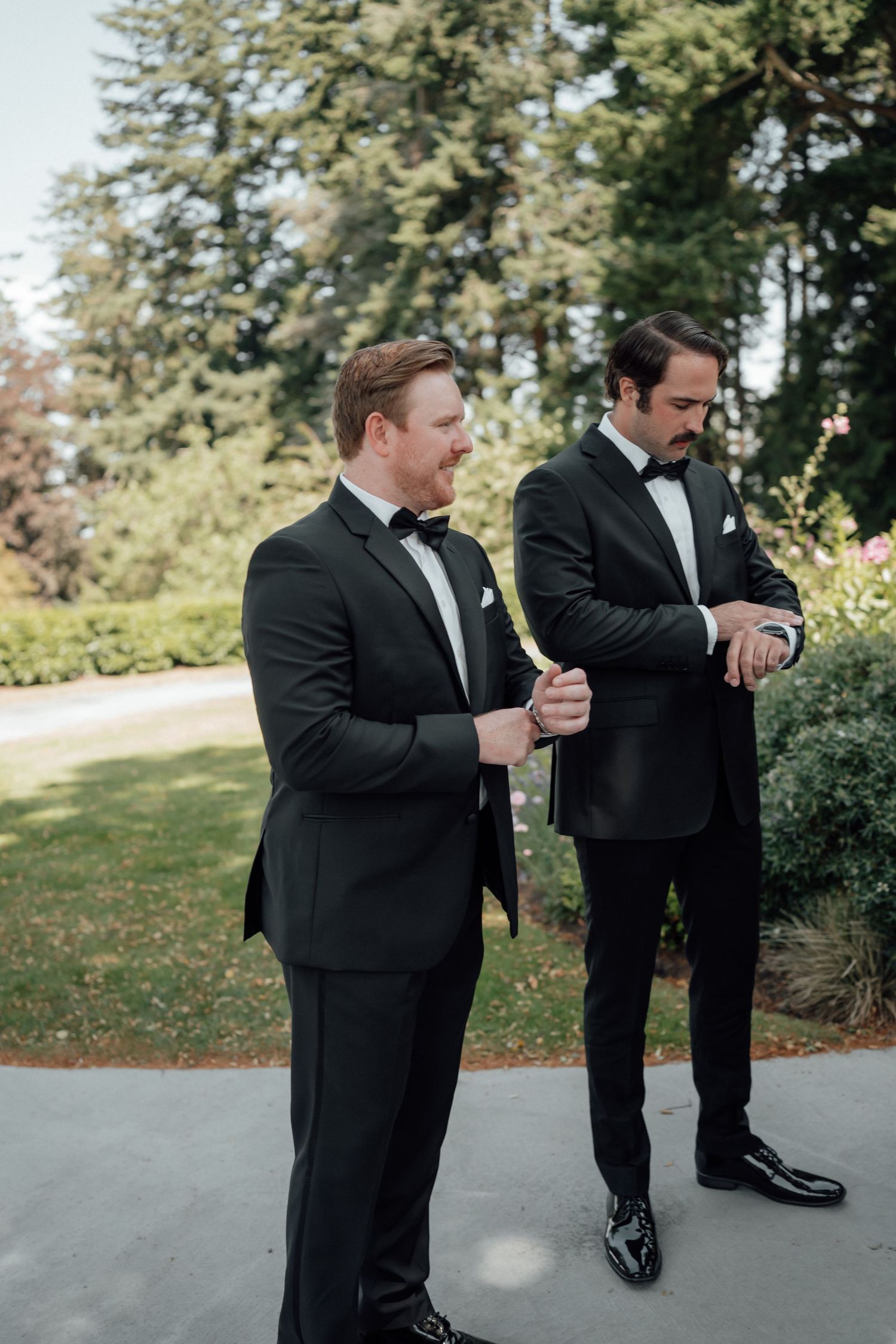 Two groomsmen in black tuxedos check their watches while standing on a pathway.