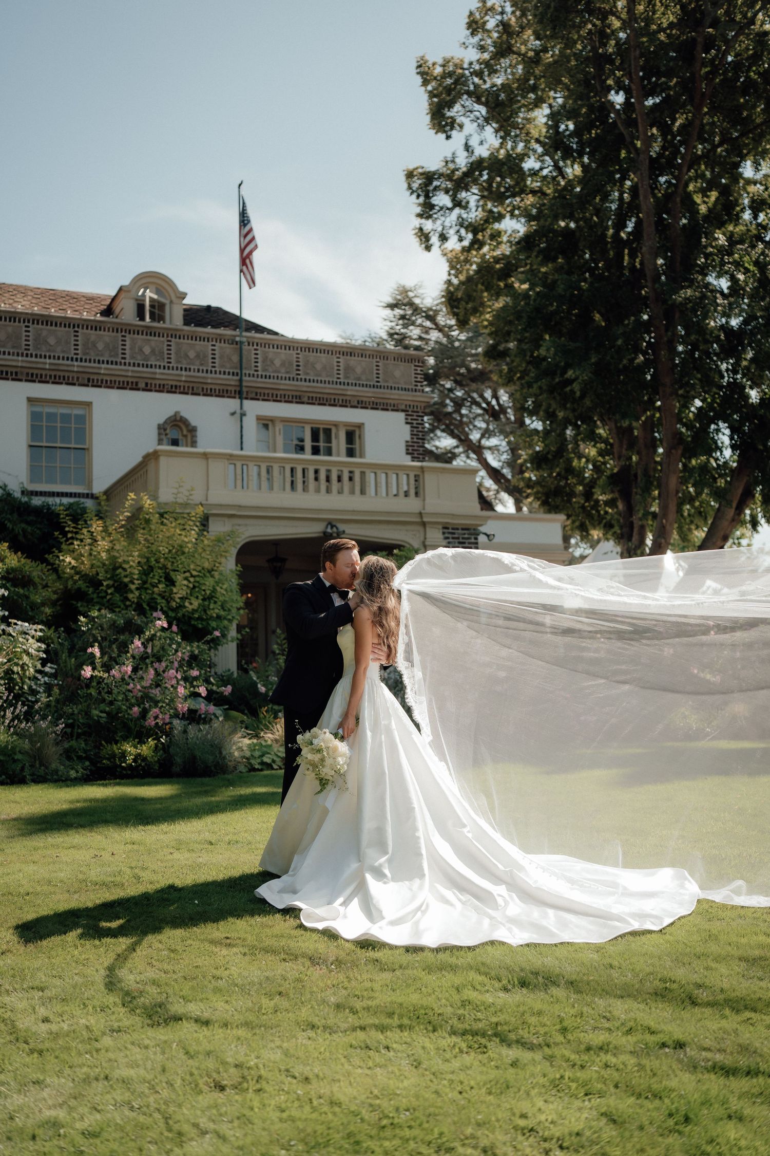 A romantic wedding portrait with a dramatic flowing veil against a stately building with an American flag.
