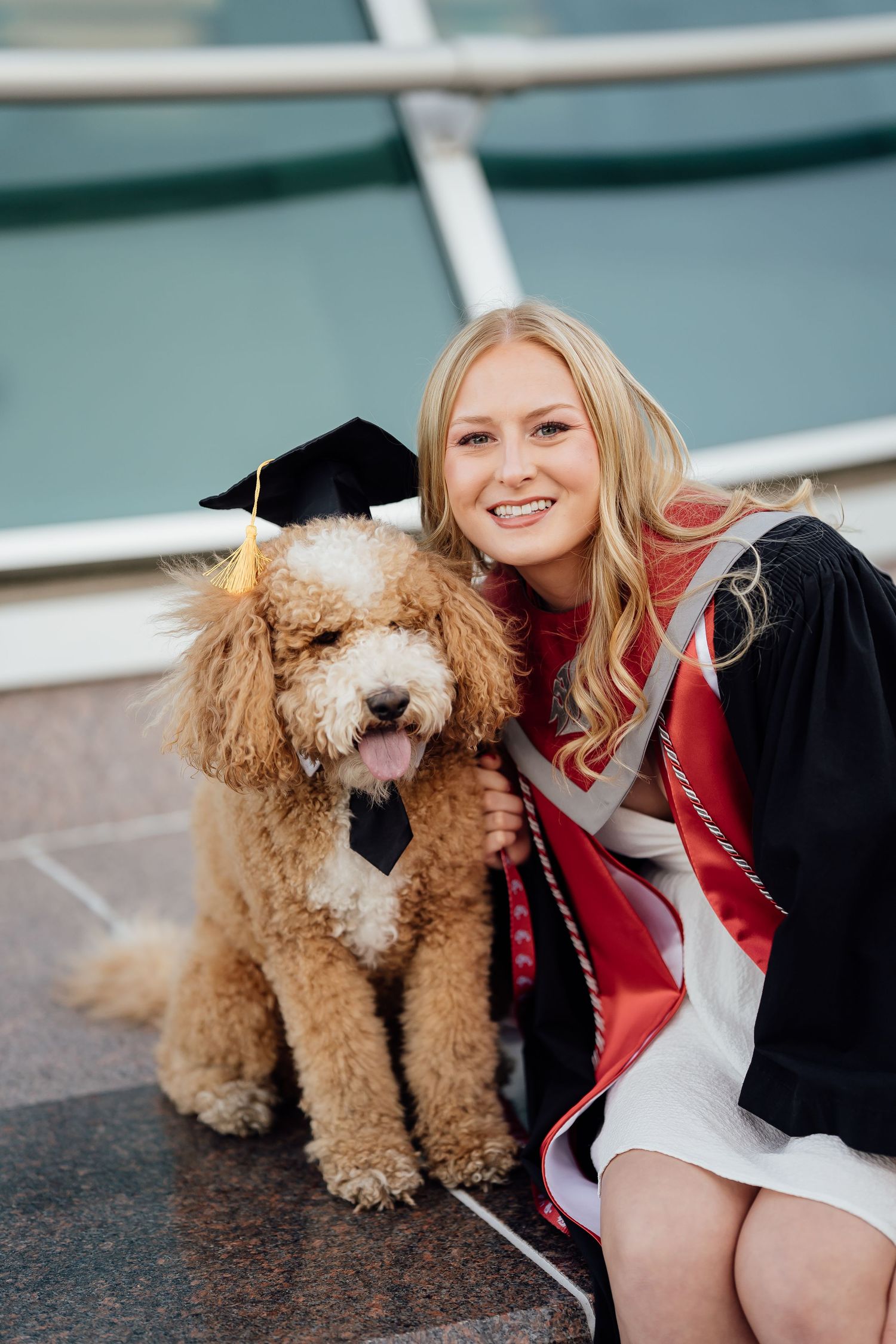 Goldendoodle dog wearing graduation cap and bowtie sits with graduate in red stole on steps.