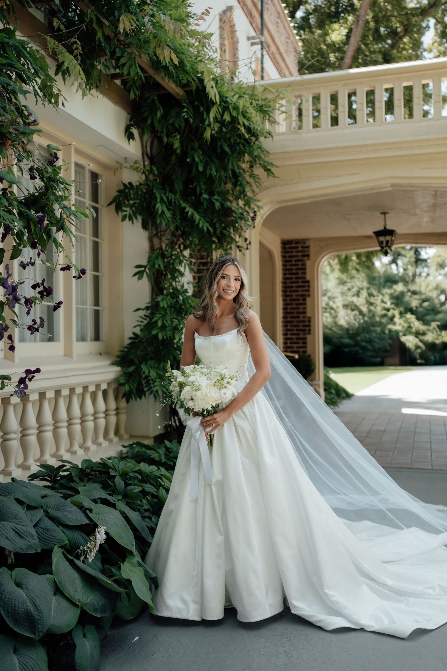 A classic bridal portrait against a white pillared mansion with lush greenery and a sweeping veil.