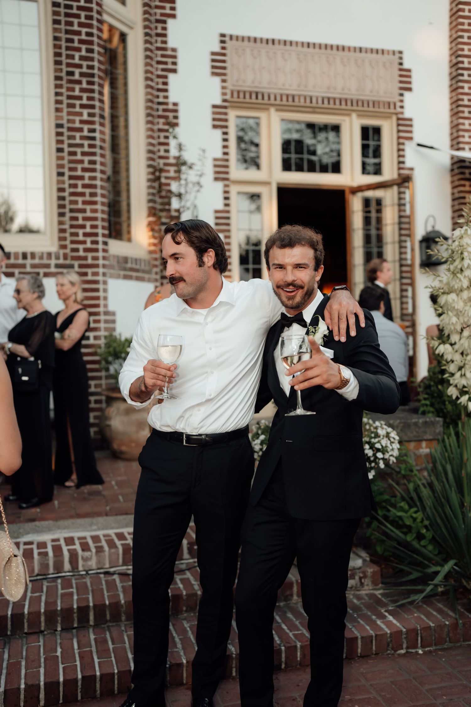 Two wedding guests in formal attire share a celebratory moment with drinks outside a brick building.