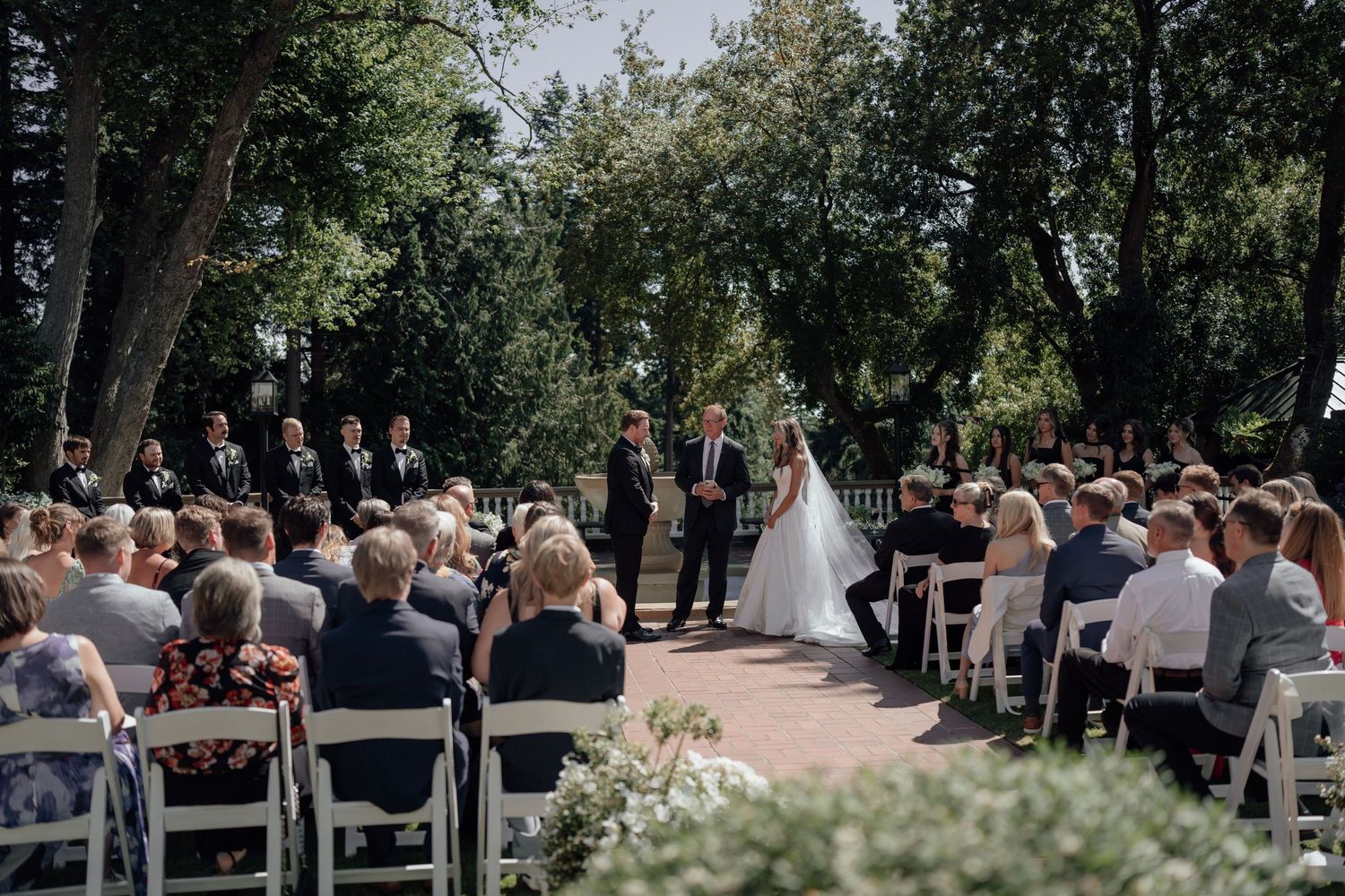 Outdoor wedding ceremony set in a lush garden with white chairs and guests seated beneath tall trees.