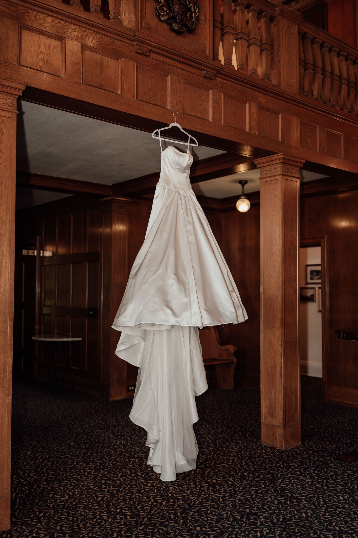 White wedding dress hanging from wooden beams in a rustic venue with dramatic lighting.