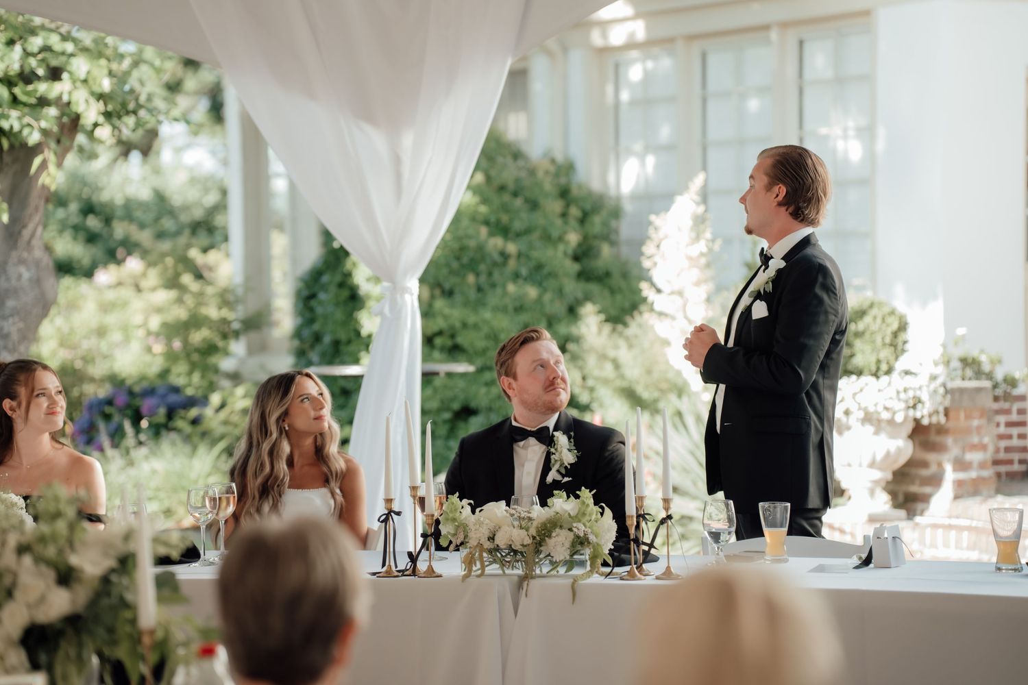 Wedding reception speech during an elegant outdoor celebration under white tent draping.