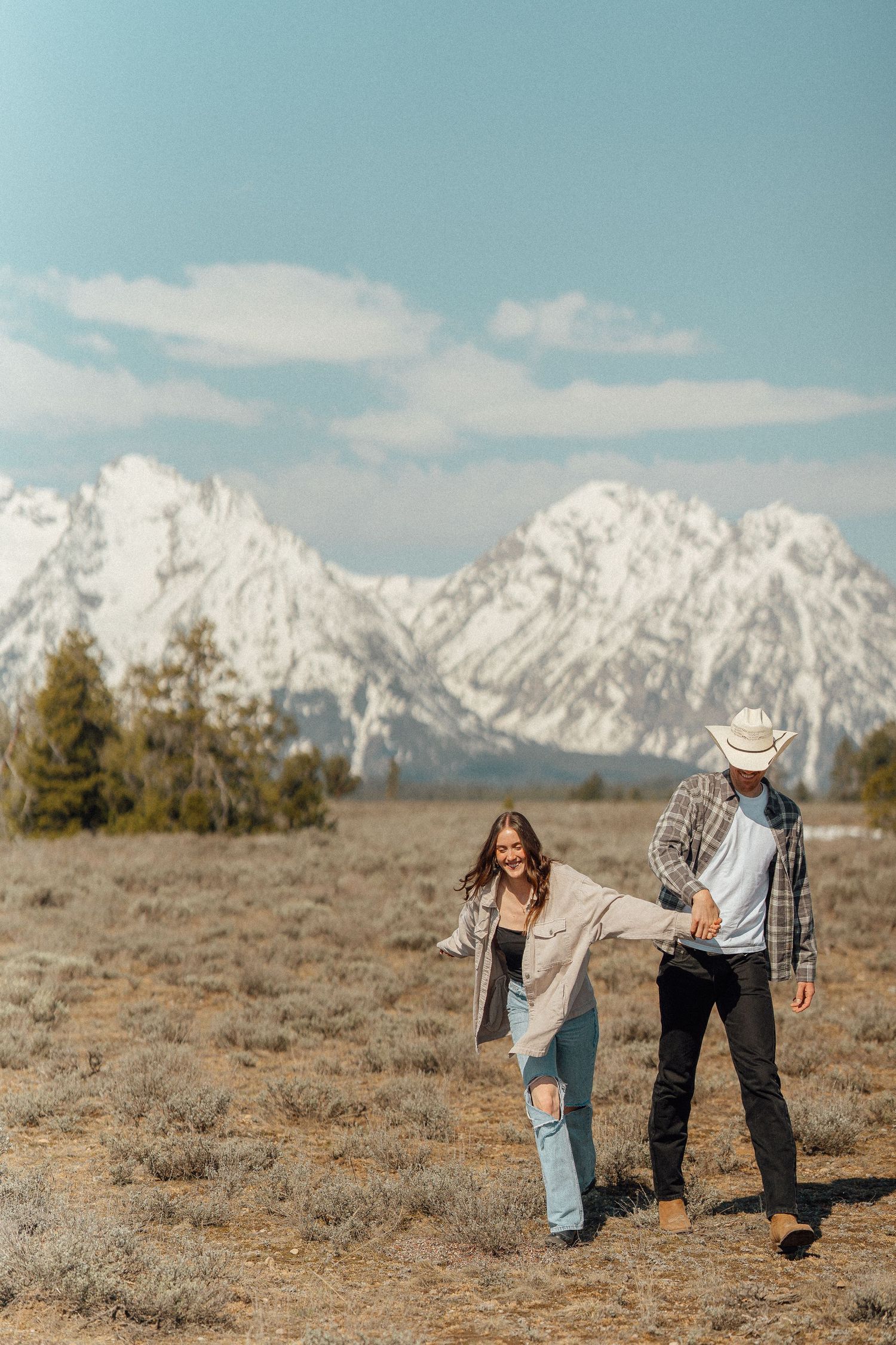 Two people walk and laugh together on an open field with snow-capped Grand Teton mountains rising majestically in the background.