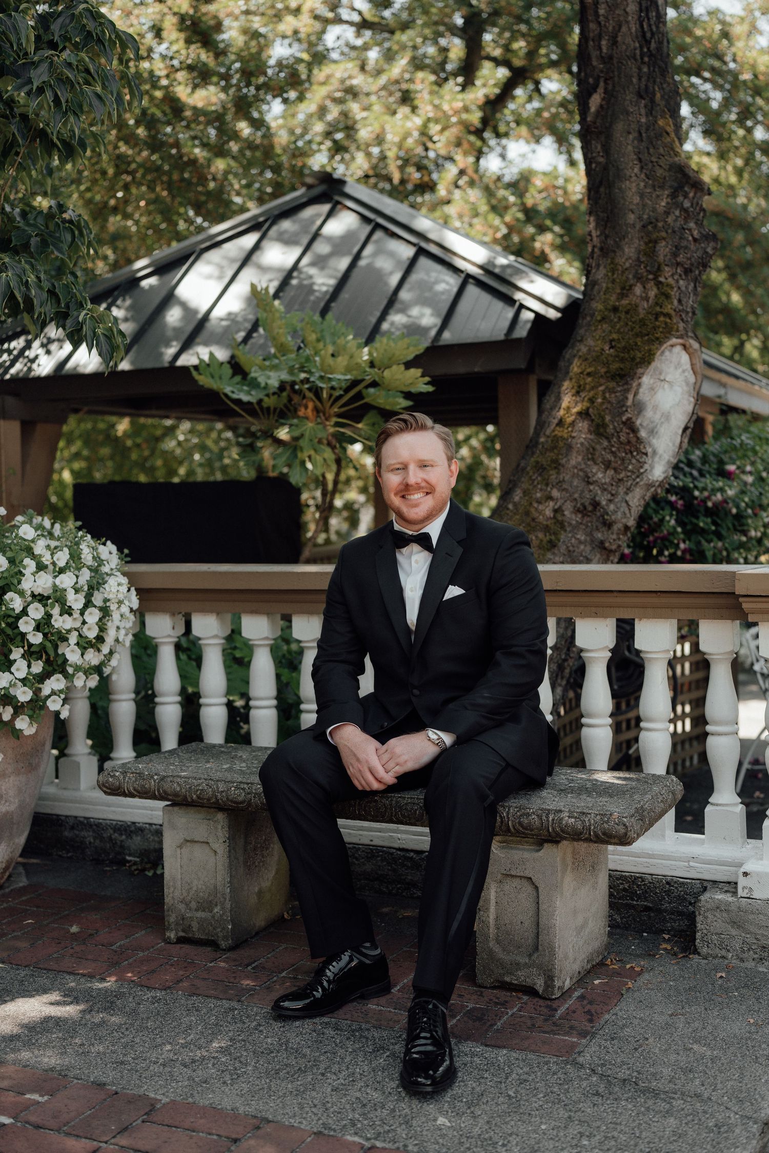 A formal portrait of a wedding attendee in a black tuxedo seated on a stone bench outdoors.
