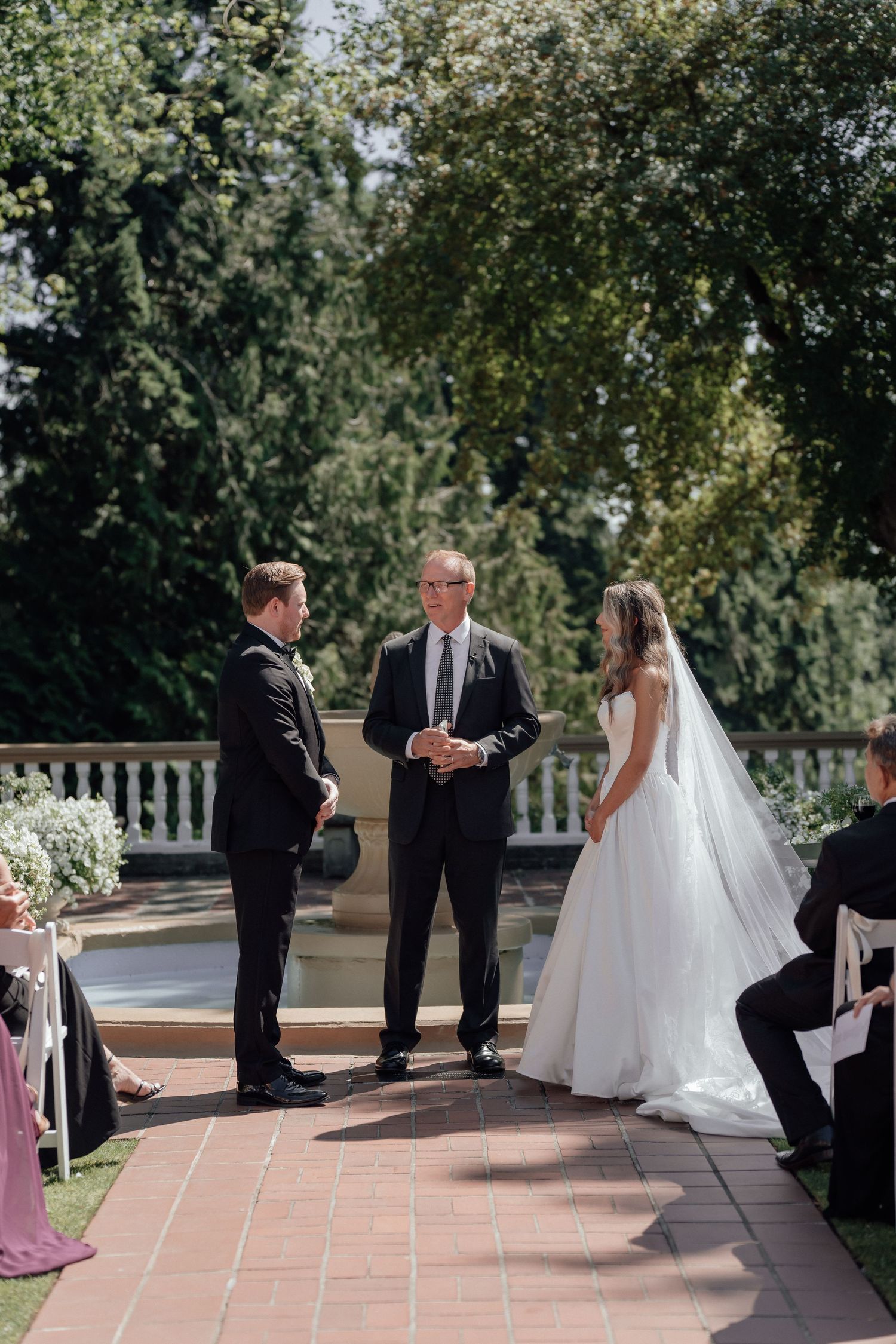 An outdoor wedding ceremony takes place on a brick patio surrounded by lush greenery and trees.