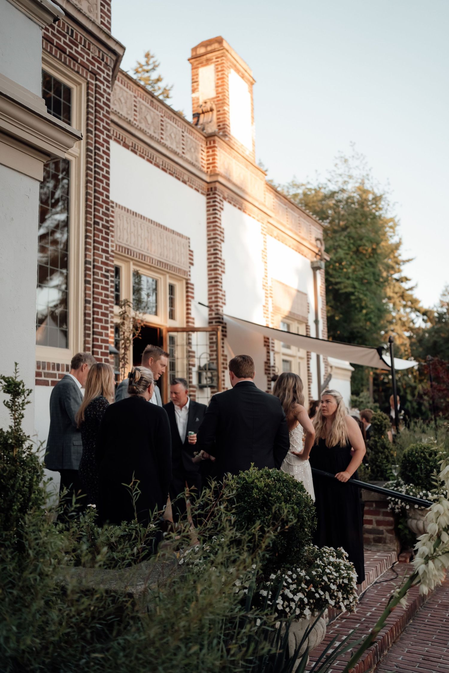 Wedding guests mingle outside a beautiful brick building with white trim during sunset.