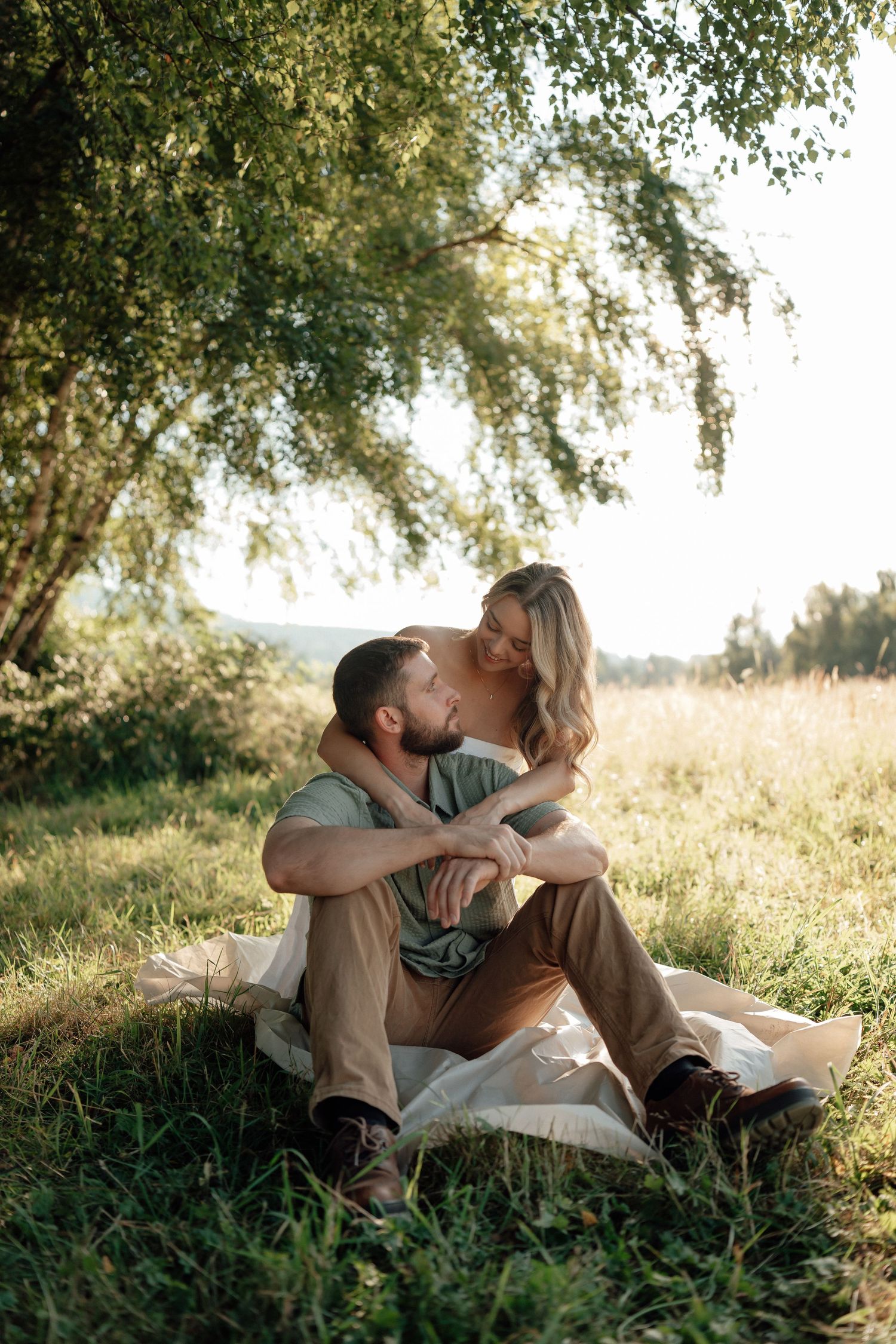 A romantic couple relaxes together on the grass in golden sunlight beneath overhanging tree branches.