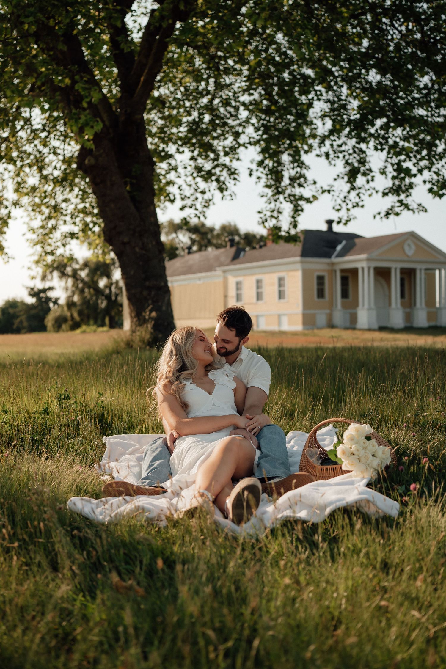 A romantic sunset picnic on a blanket in front of a historic white plantation-style house with a tree overhead.
