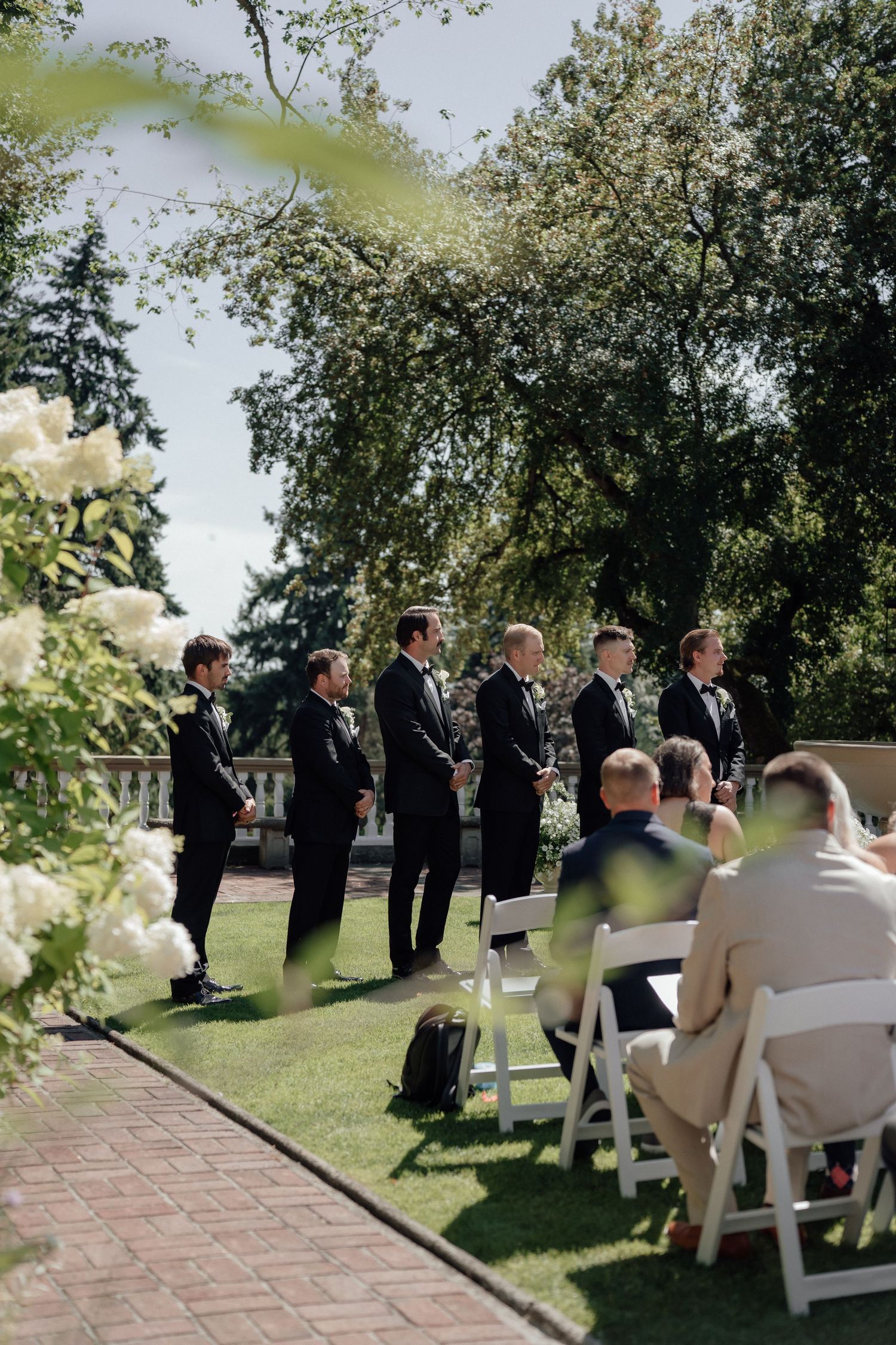 Groomsmen in black tuxedos line up outdoors during a wedding ceremony with guests seated on white chairs.