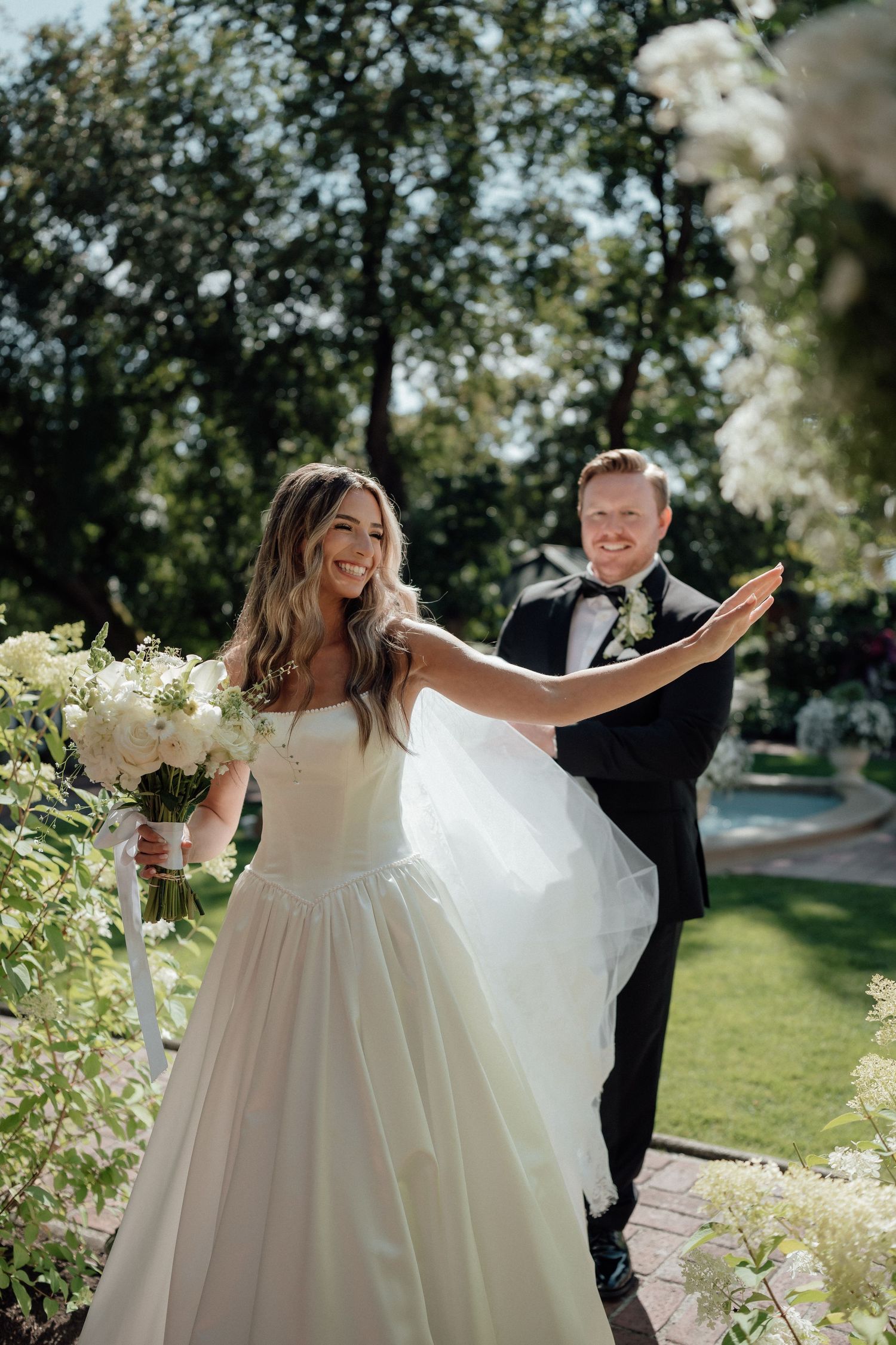 A joyful wedding moment captured outdoors with the bride in a flowing white gown holding a bouquet amid greenery.
