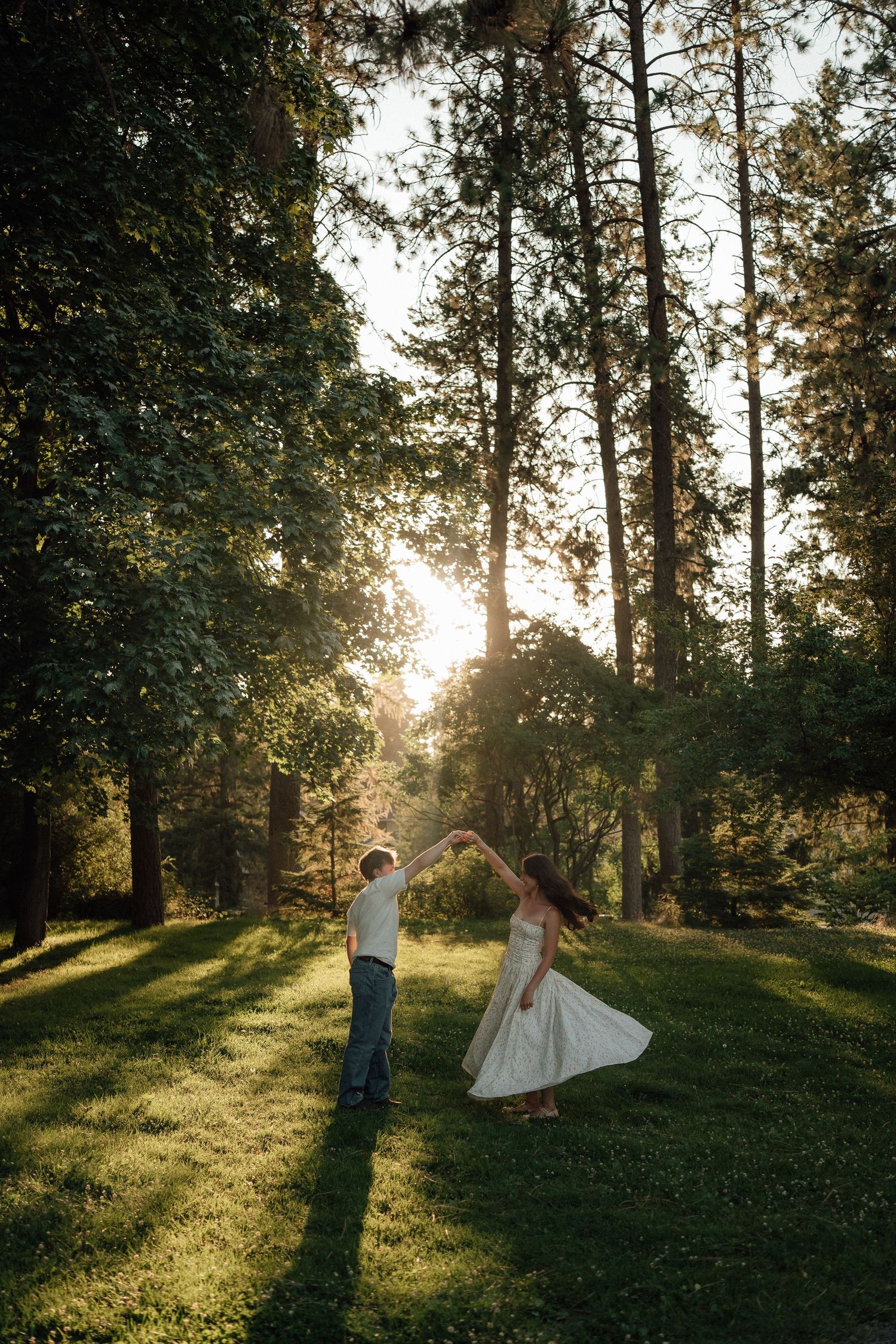 A couple dances together in a sunlit wooded clearing during golden hour creating dreamy silhouettes against tall trees.