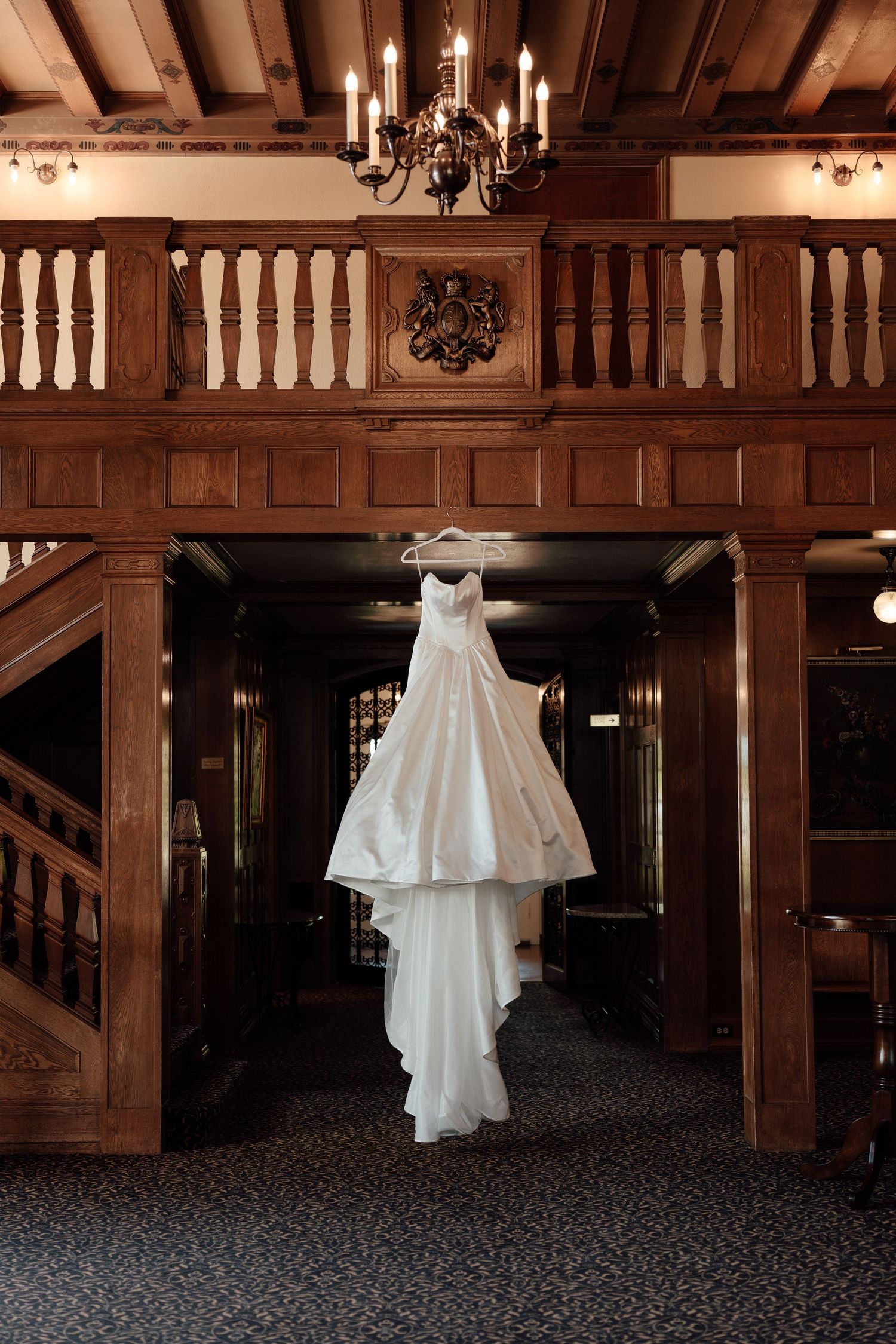 An elegant white wedding dress hangs from a chandelier in a wooden-paneled hallway with ornate architectural details.