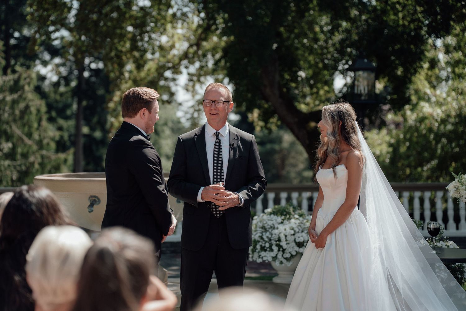 An outdoor wedding ceremony in a garden setting with an officiant standing between a bride and groom.
