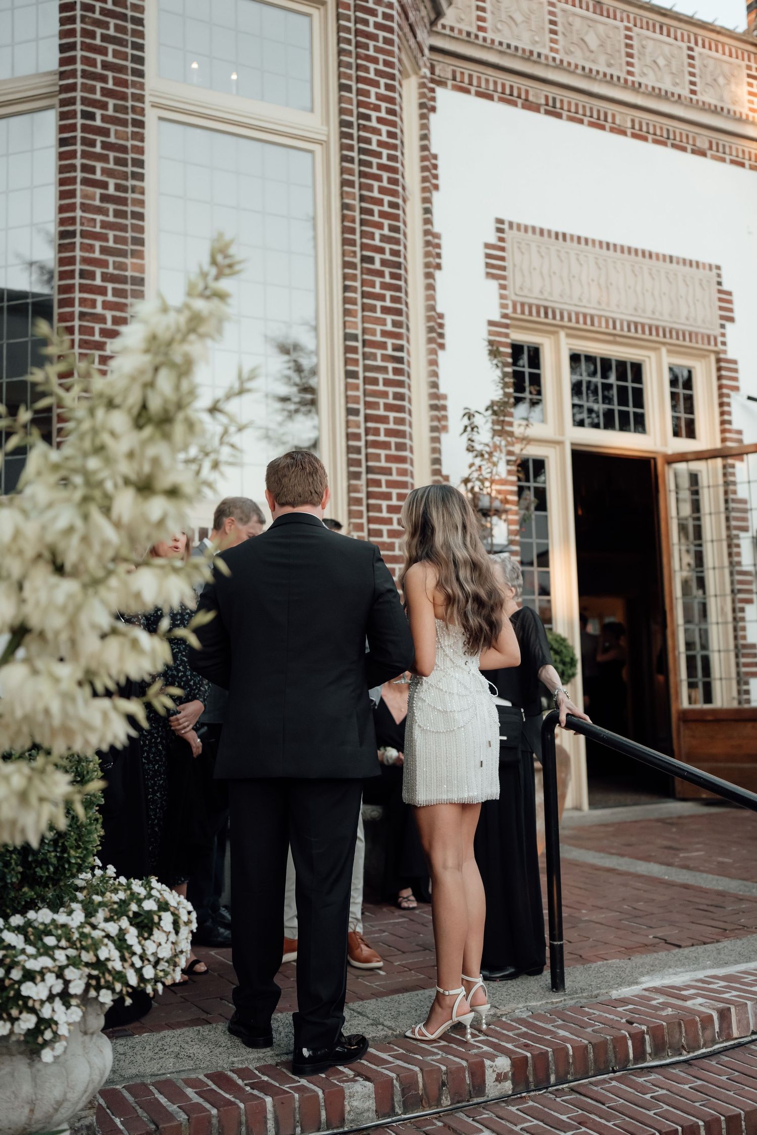 Wedding guests gather outside a historic brick building entrance decorated with white floral arrangements.