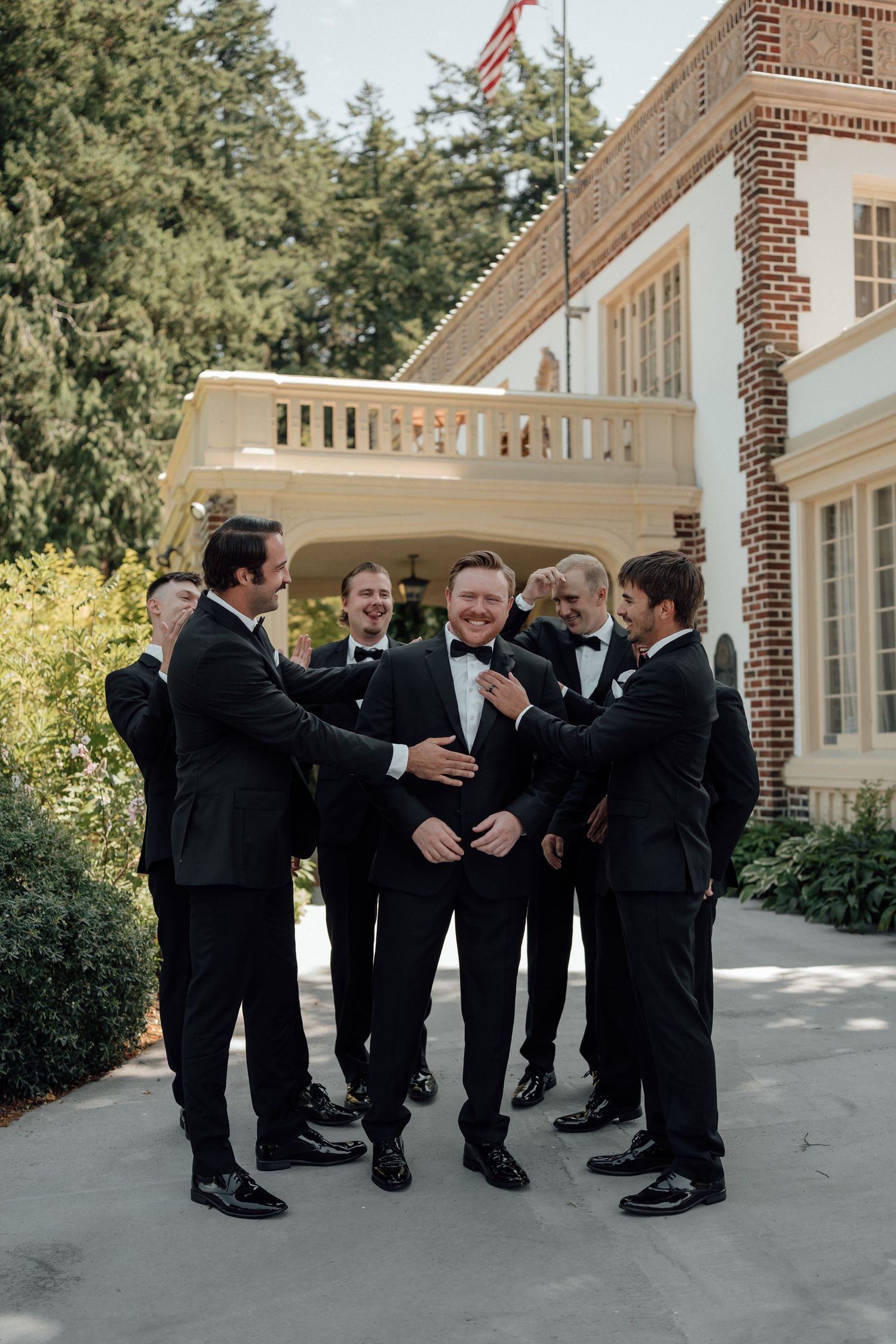 A groomsmen group in black tuxedos share a lighthearted moment outside a brick building with an American flag.