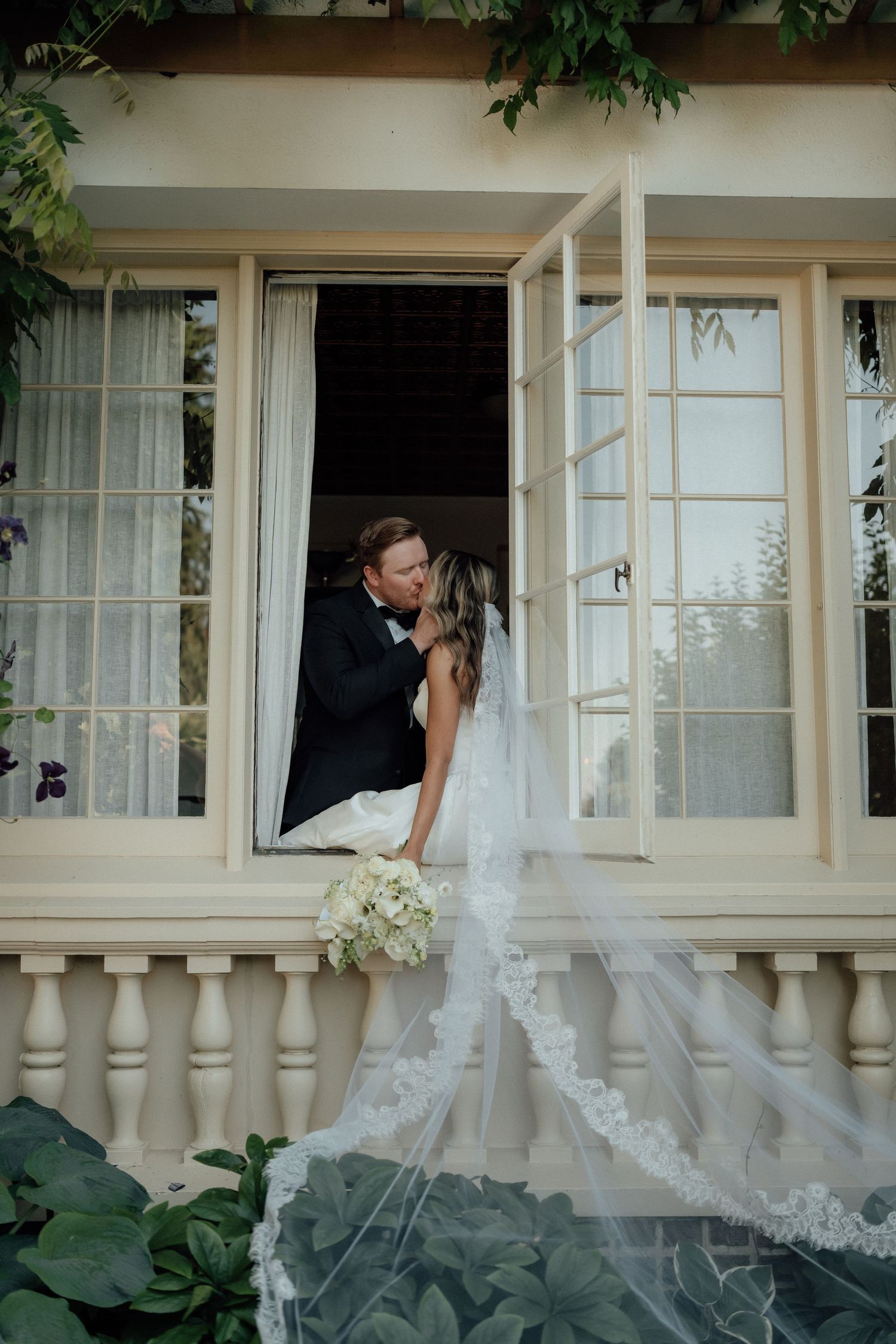 An intimate wedding moment captured on an elegant white balcony with trailing vines and french doors.