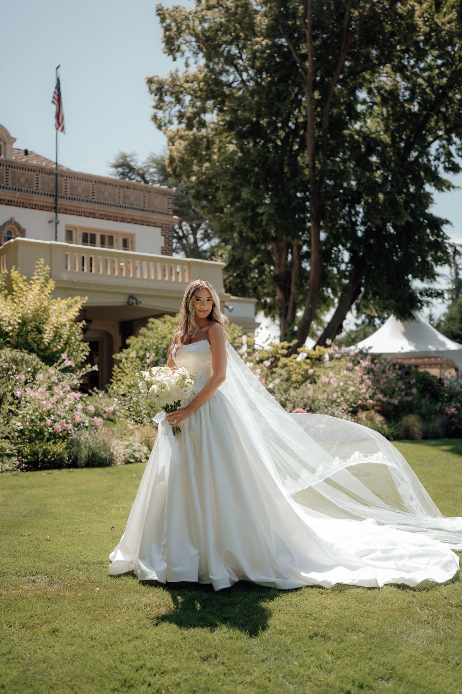 Elegant white wedding gown with flowing train photographed on manicured lawn of historic building.