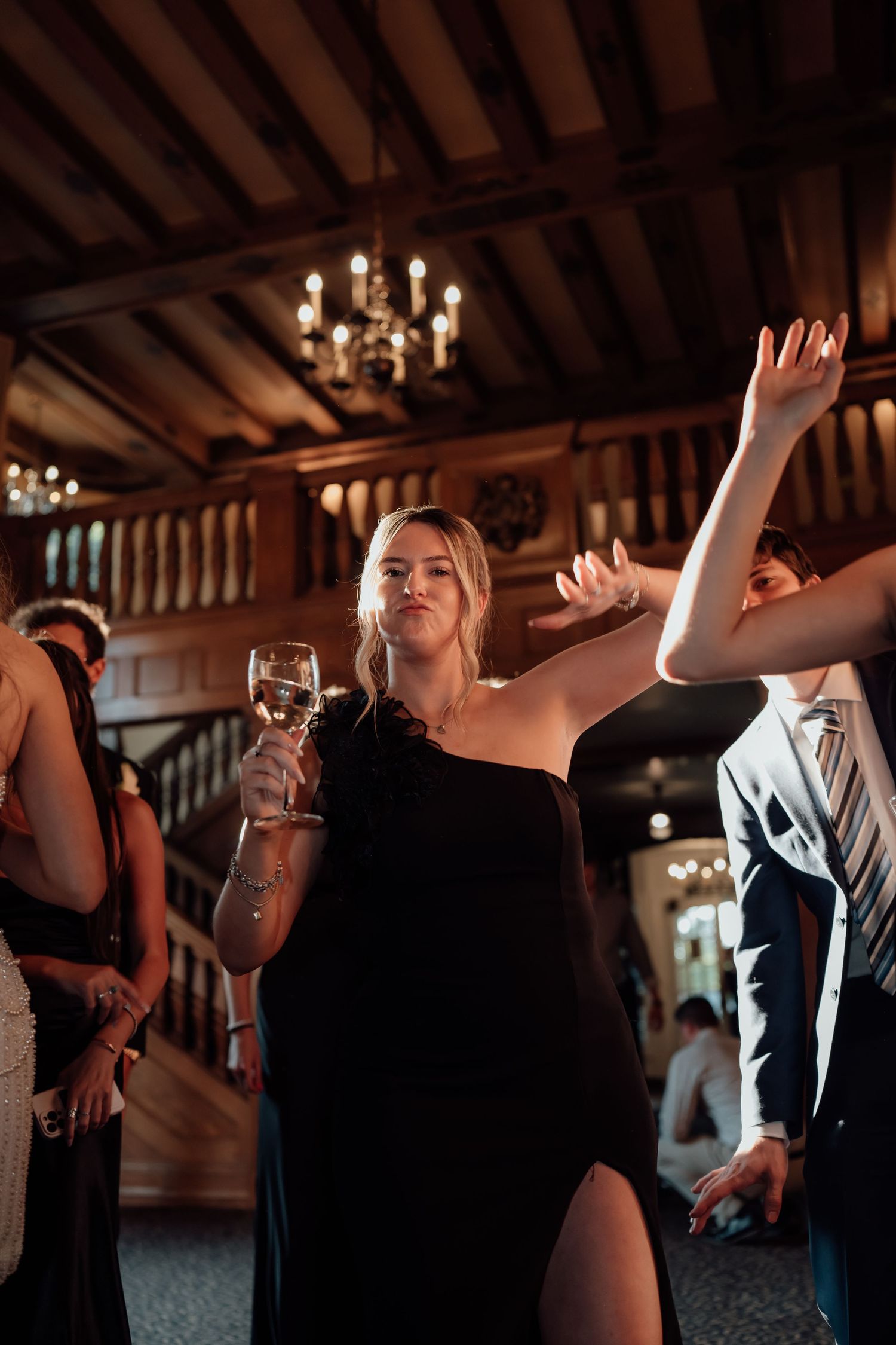 A wedding guest in a black dress dances joyfully with raised arms at an indoor reception.