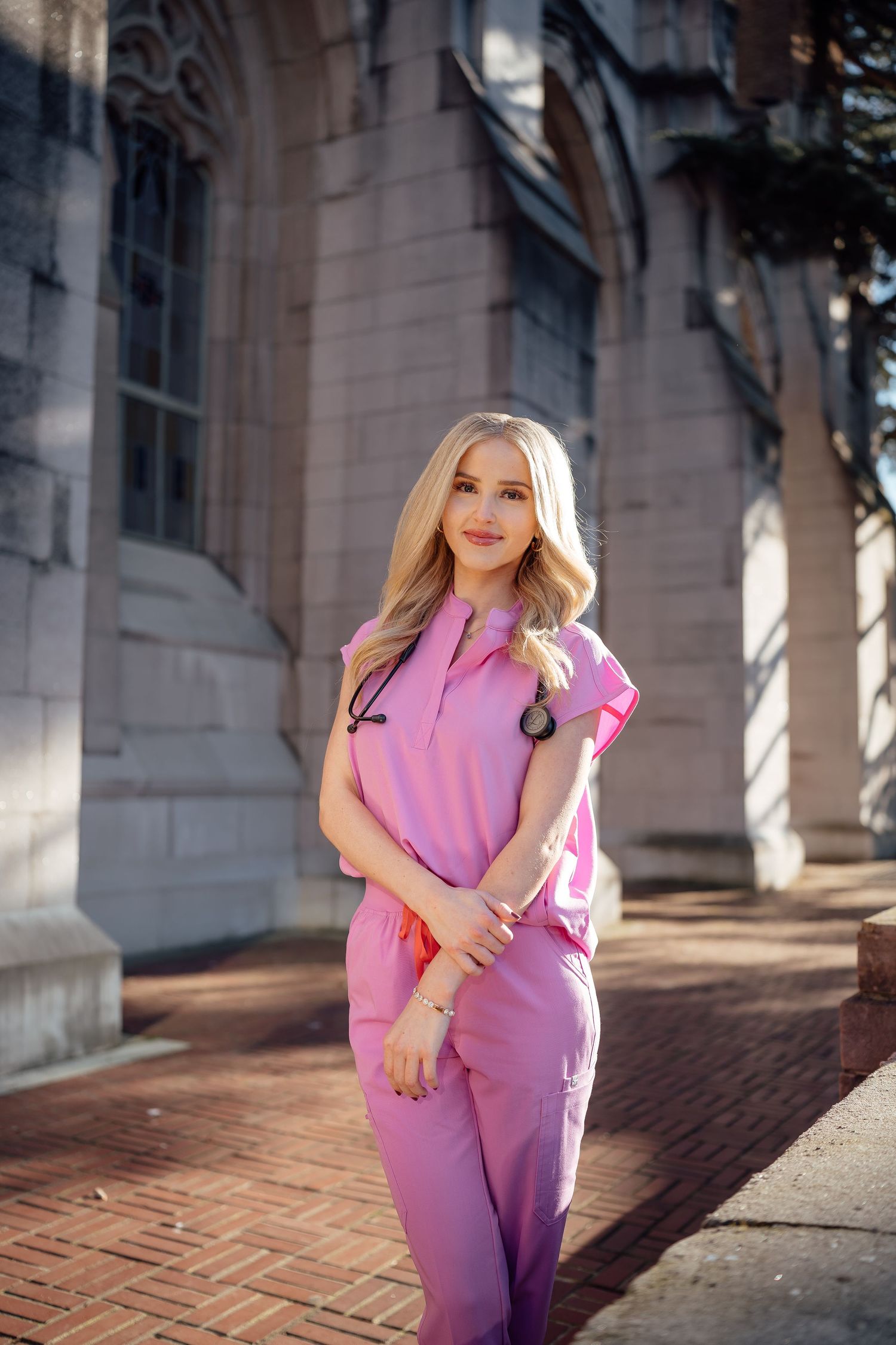 A person in a lavender scrub uniform poses outdoors against a brick and stone building in natural lighting.