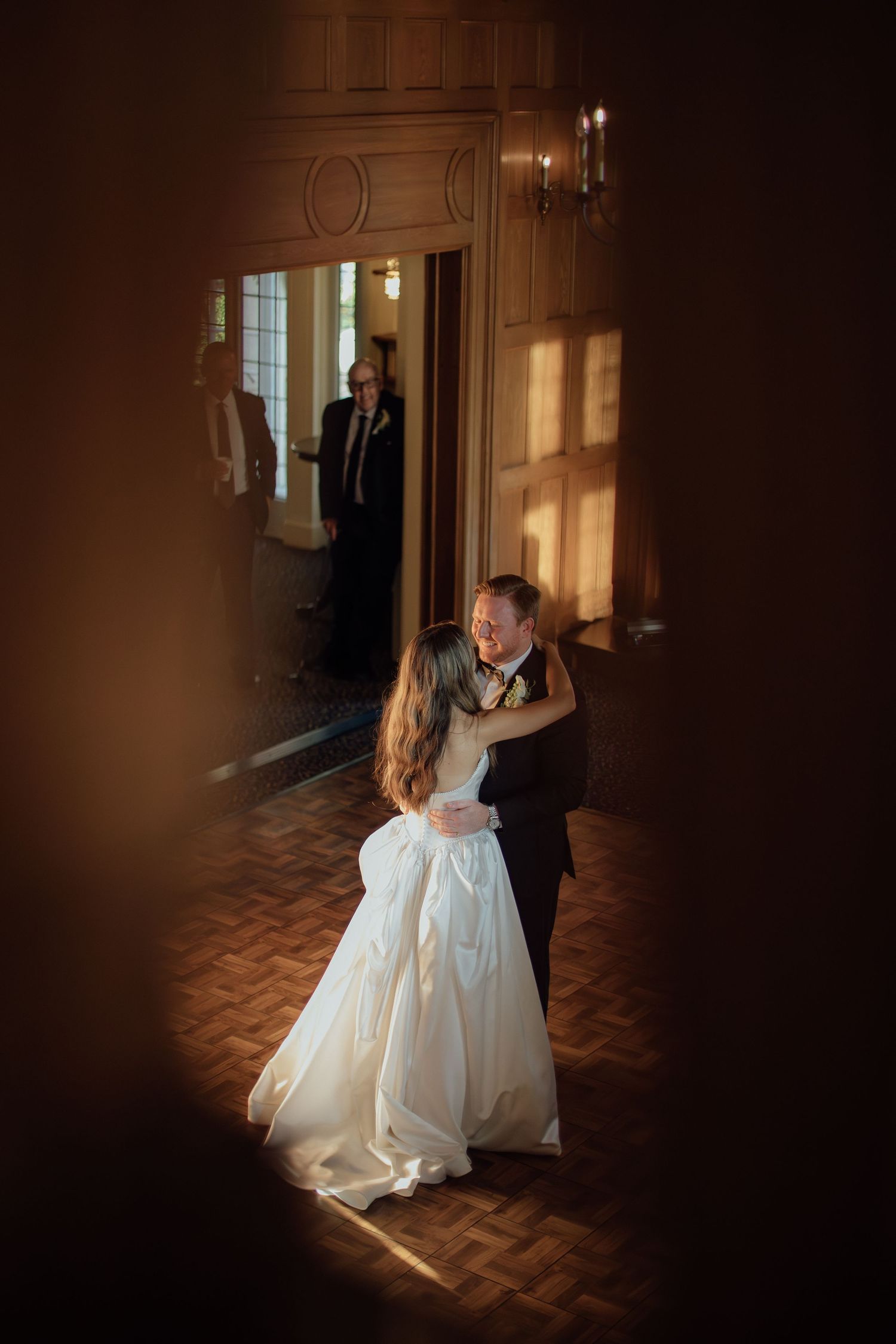A romantic first dance captured in dramatic lighting through a doorway on a parquet floor.