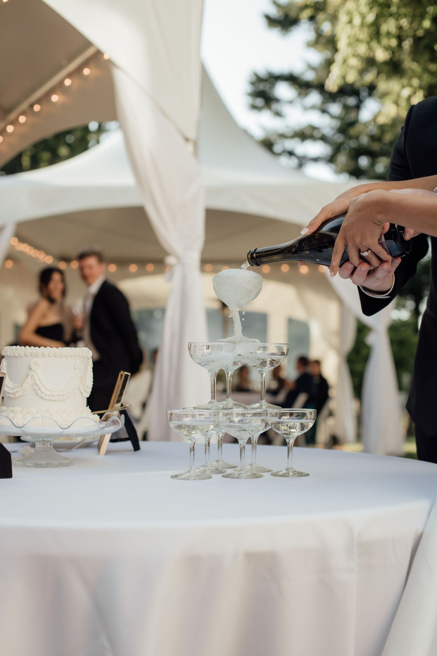 Champagne being poured into a tower of glasses at an elegant outdoor wedding reception with twinkling lights.