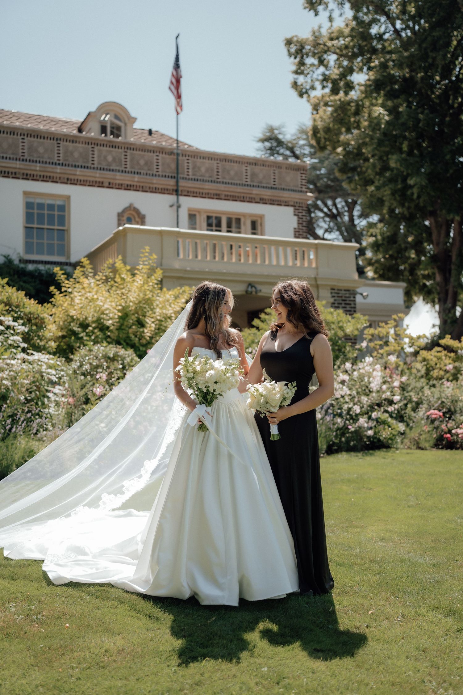 A bride in a flowing white gown stands with a person in black beside a classic white building with an American flag.